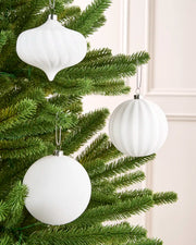 Three white ribbed Christmas tree ornaments, including two spheres and one teardrop shape, hanging from a green artificial Christmas tree against a light cream paneled wall.