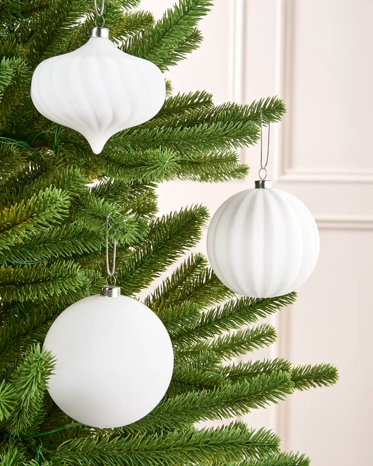 Three white ribbed Christmas tree ornaments, including two spheres and one teardrop shape, hanging from a green artificial Christmas tree against a light cream paneled wall.