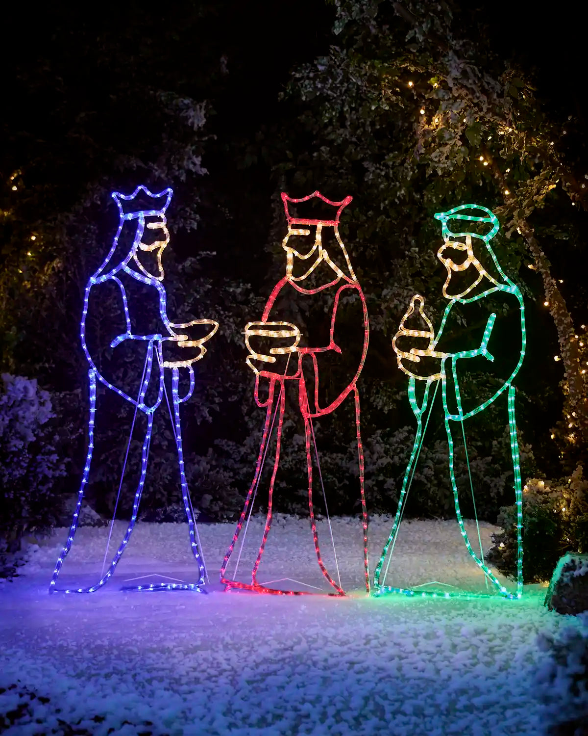Three wise men in blue, red and green silhouette on snowy grass against a garden fence and trees in background 