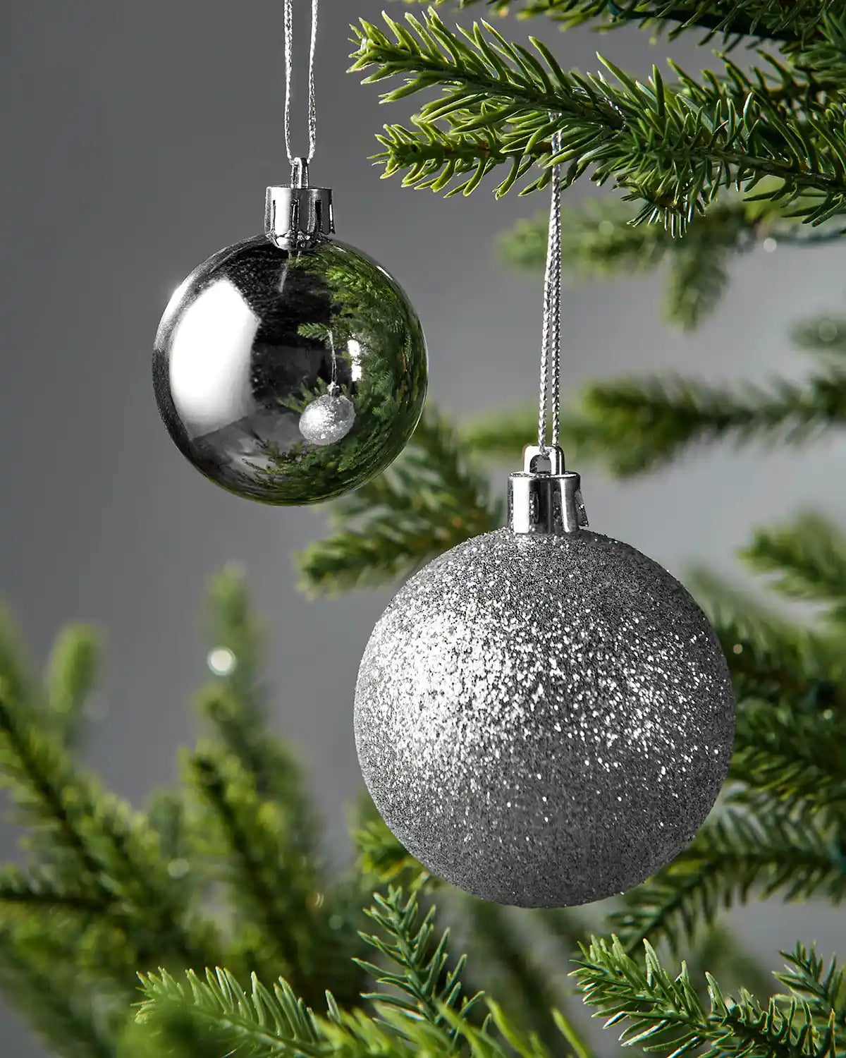 Two Christmas ornaments, one gloss silver and one silver glittery, hanging on a tree branch.