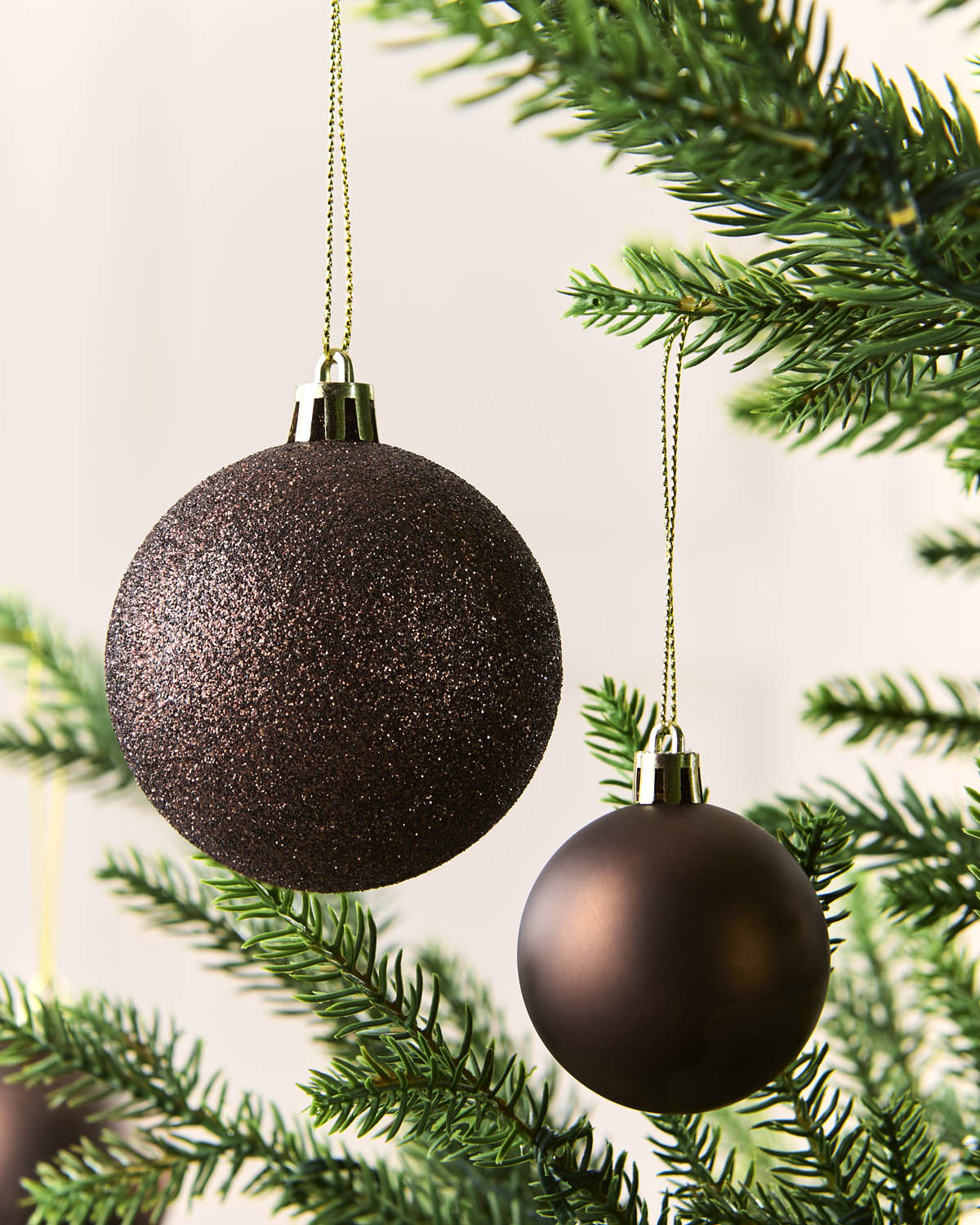 Two Espresso Christmas tree ornaments hanging from green pine branches against a soft off-white background.