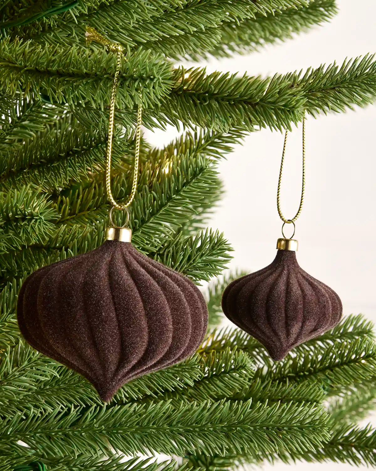 Two Espresso coloured velvet Christmas tree ornaments hanging from gold chains on green pine tree branches against a light background.