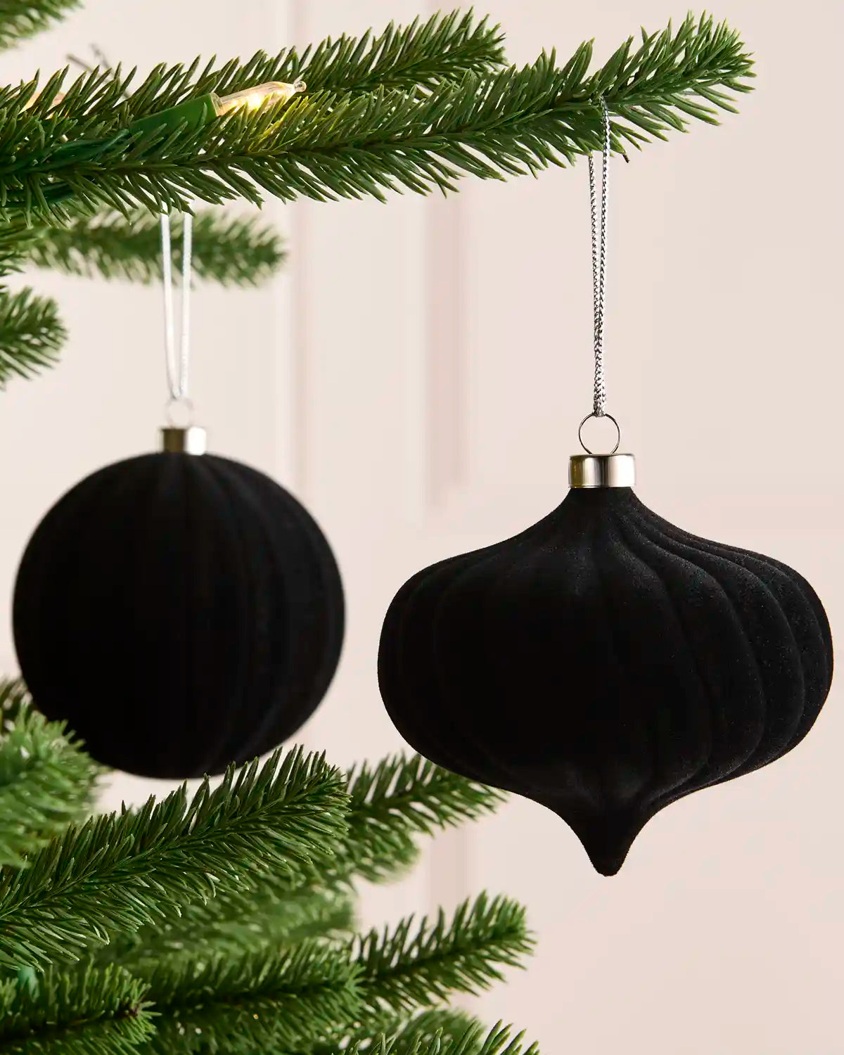 Two black Christmas tree ornaments hanging from green pine branches against a soft beige background.