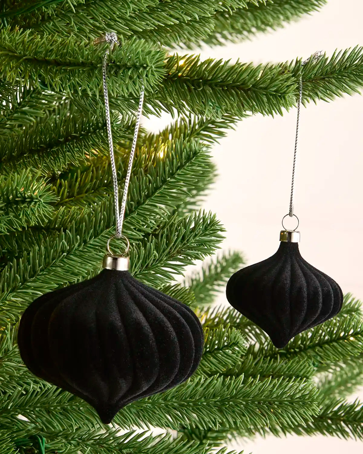 Two black ribbed Christmas tree ornaments hanging from silver strings on green pine branches against a light cream background.