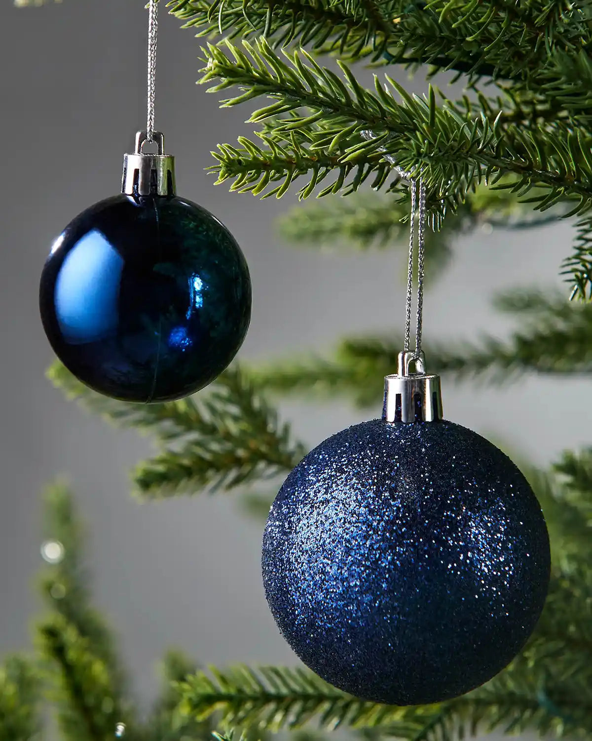 Two blue Christmas baubles hanging on a tree branch with a grey blurry background