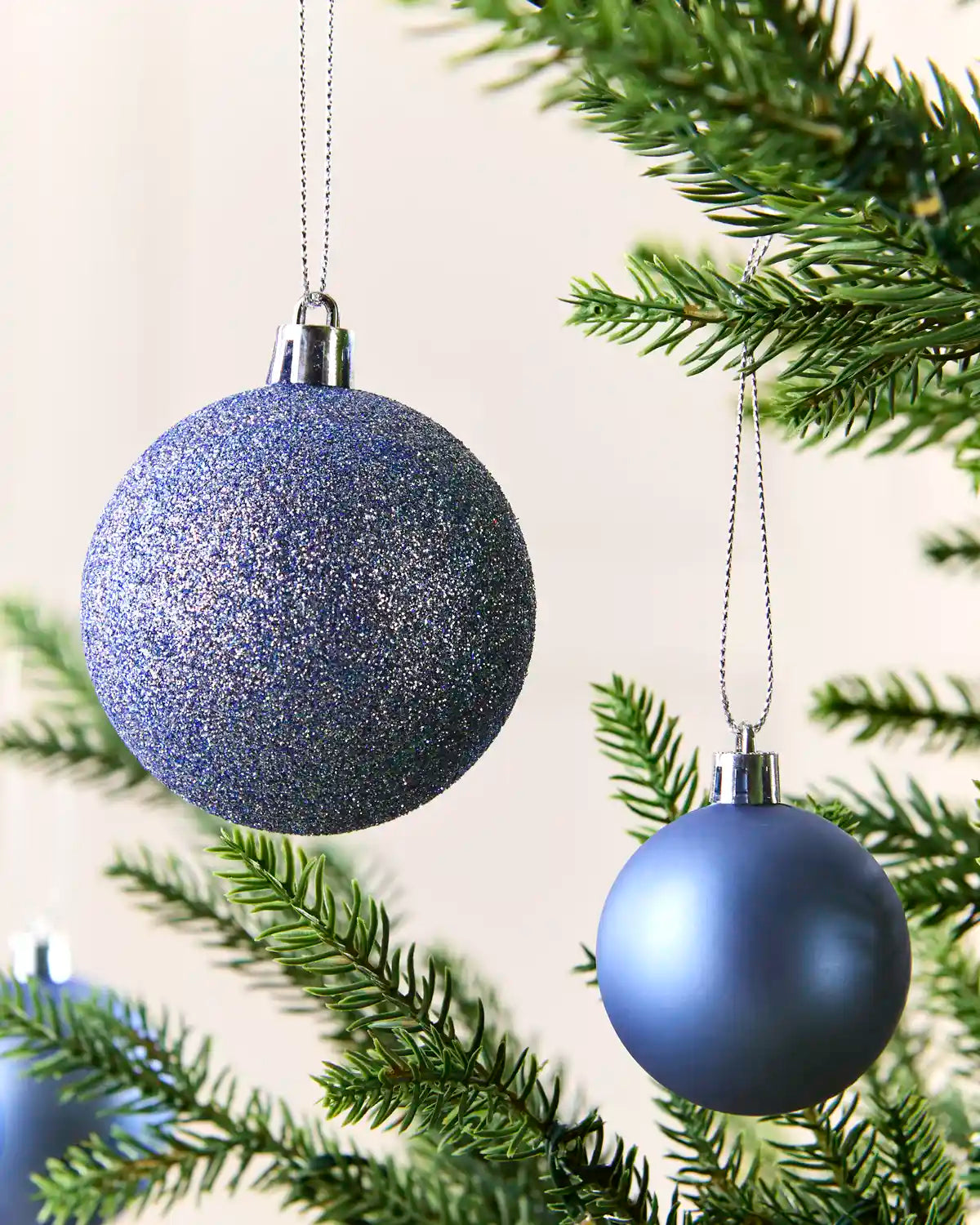 Two blue Christmas tree ornaments hanging from green pine branches against a soft cream background.