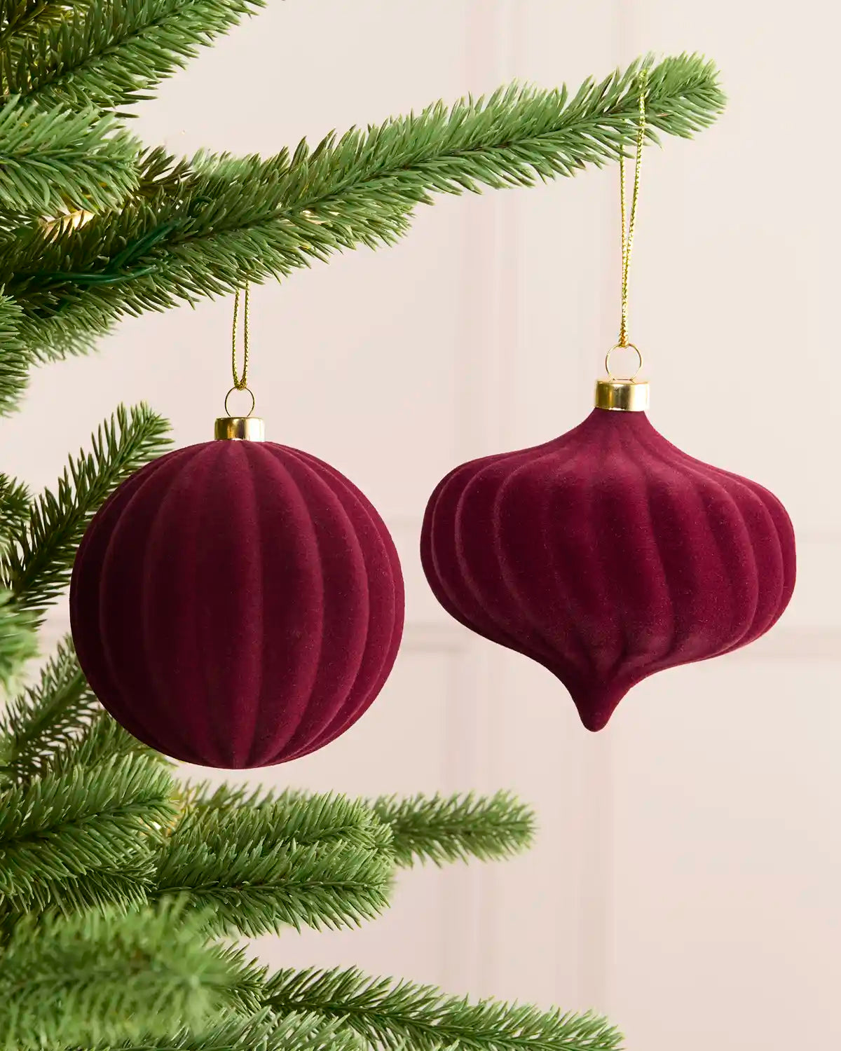 Two burgundy velvet Christmas tree ornaments hanging from green pine branches against a soft cream background.
