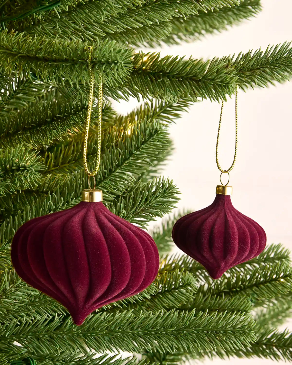 Two burgundy velvet Christmas tree ornaments with gold caps and gold hanging strings, displayed on green pine tree branches against a light background.