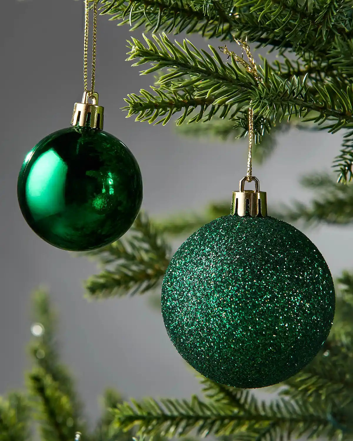 Two close up green Christmas baubles hanging on a tree branch against a grey background