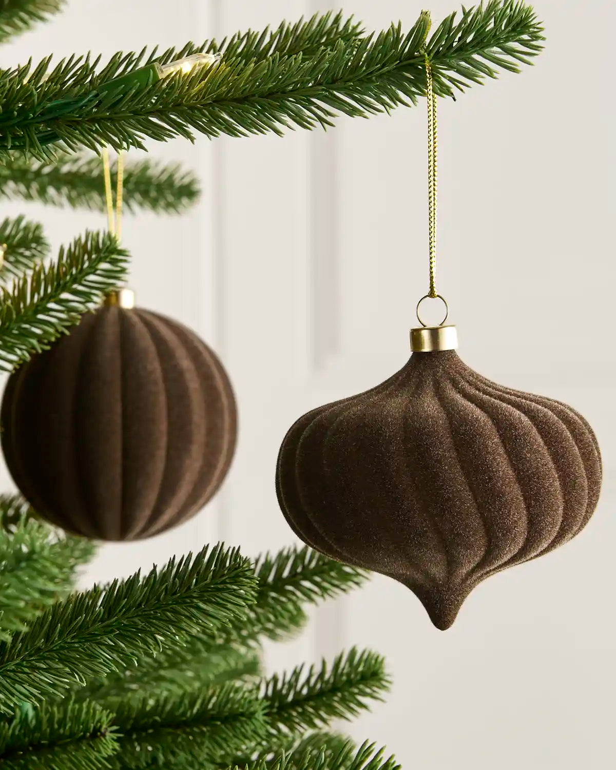 Two dark brown ribbed felt Christmas ornaments hanging from a green Christmas tree branch against a soft off-white background.