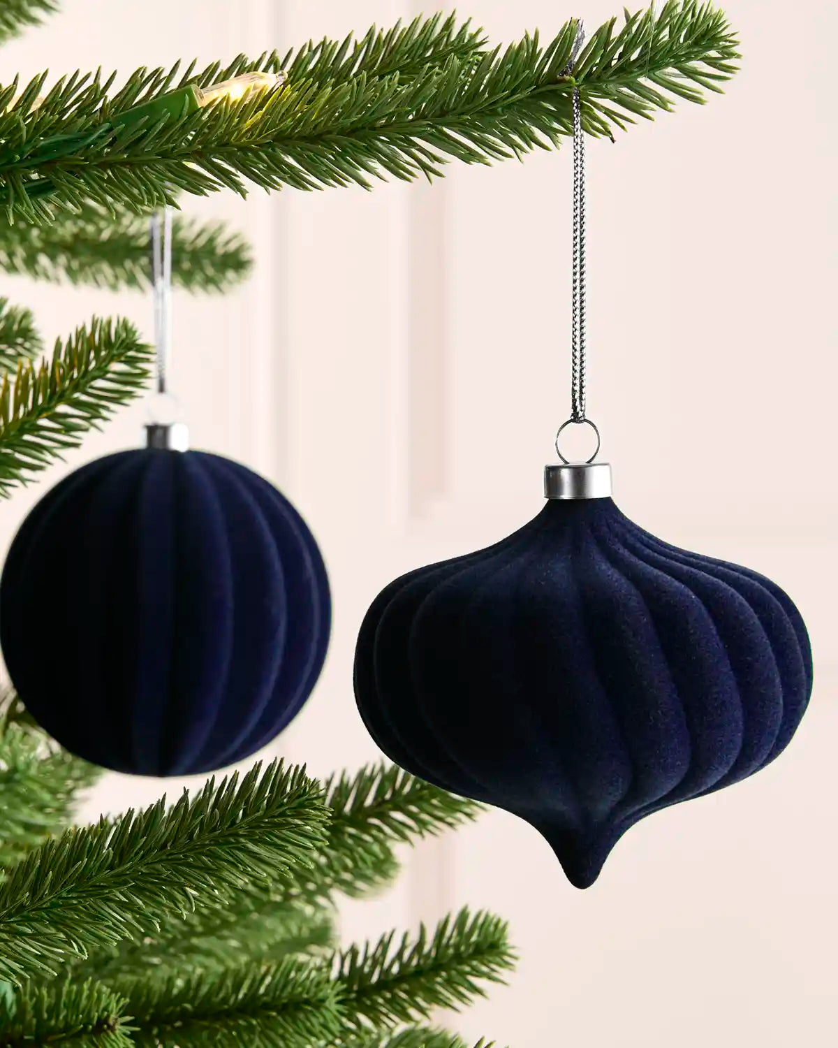 Two dark navy blue velvet Christmas tree ornaments hanging from a green pine tree branch against a soft cream background.