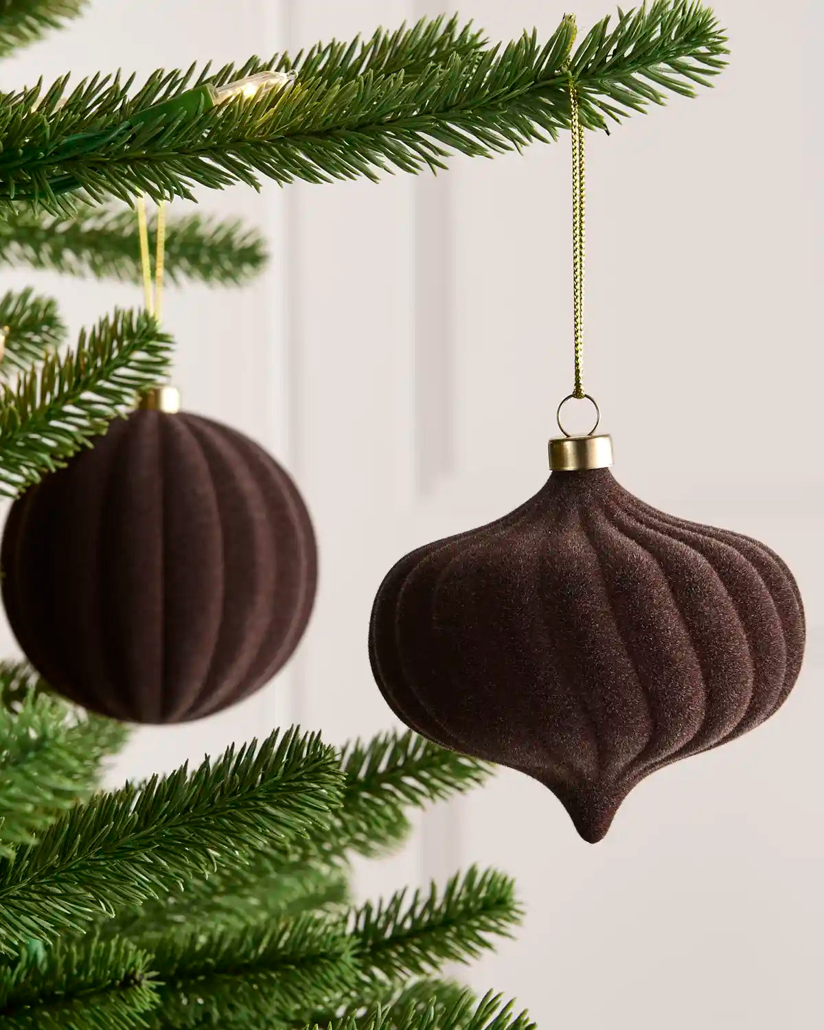 Two espresso coloured ribbed velvet Christmas tree ornaments hanging from a green pine tree branch against a light neutral background.