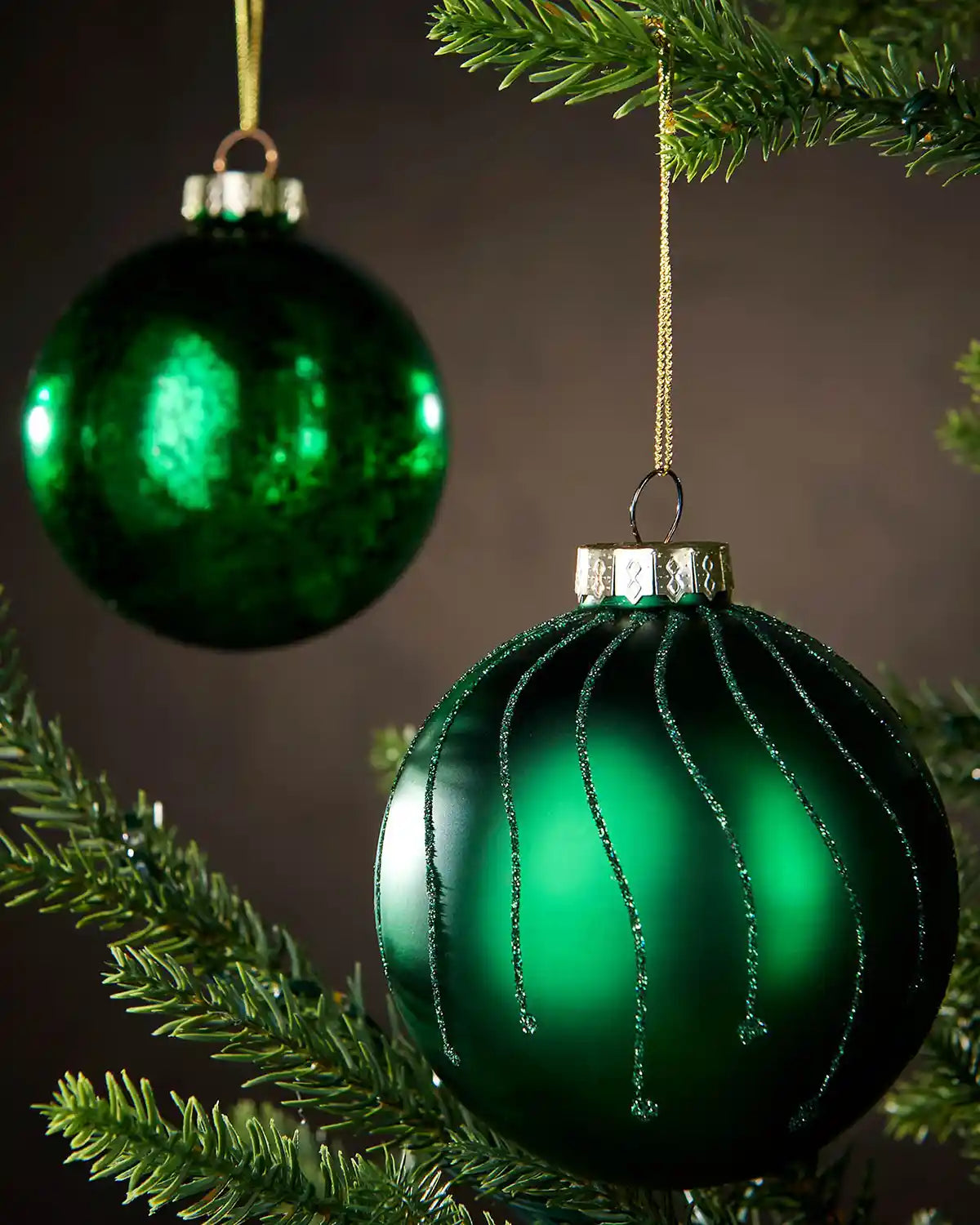 Two green glass ornaments hanging on a Christmas tree branch with a dark background.