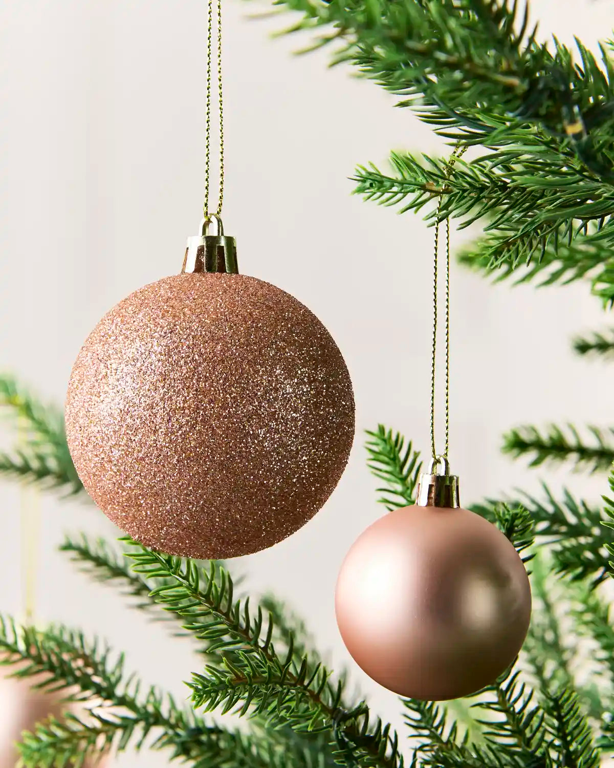 Two hanging Christmas ornaments on a tree branch: one large glittery rose-gold sphere and one smaller smooth rose-gold sphere, with green pine needles around them.