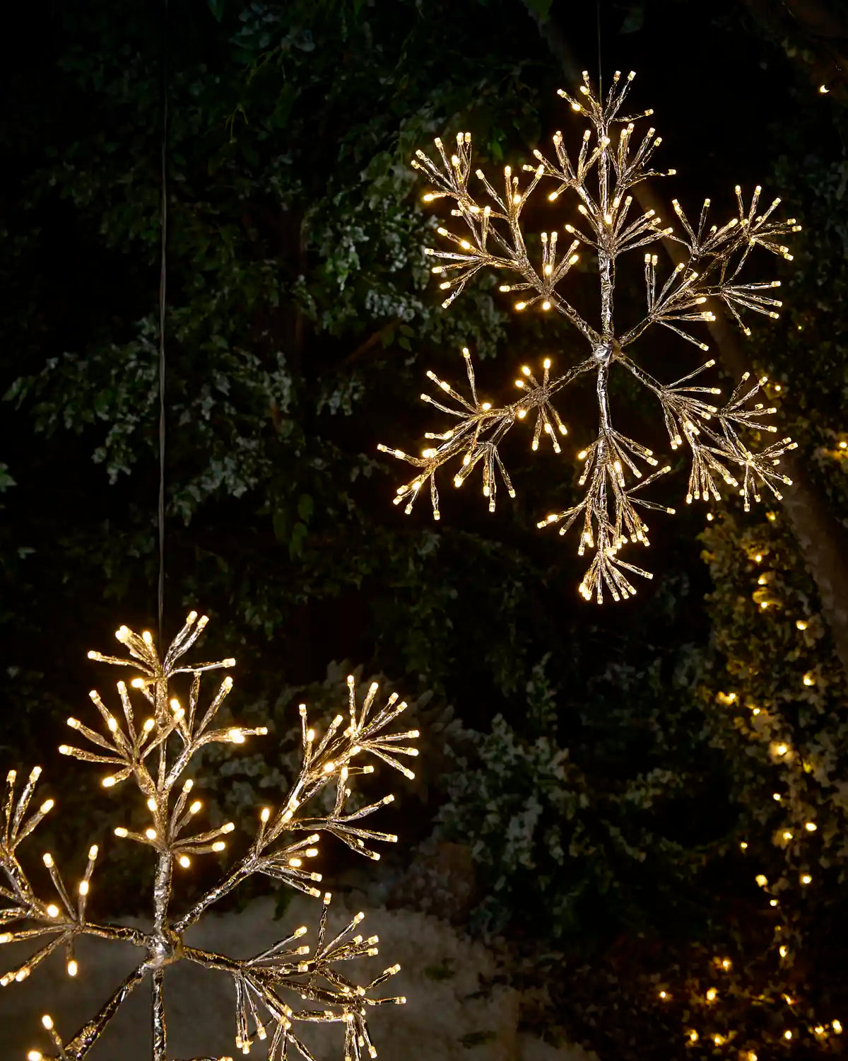 Two large hanging snowflake light decorations with warm white branch-like lights, photographed outdoors at night against dark trees and foliage.