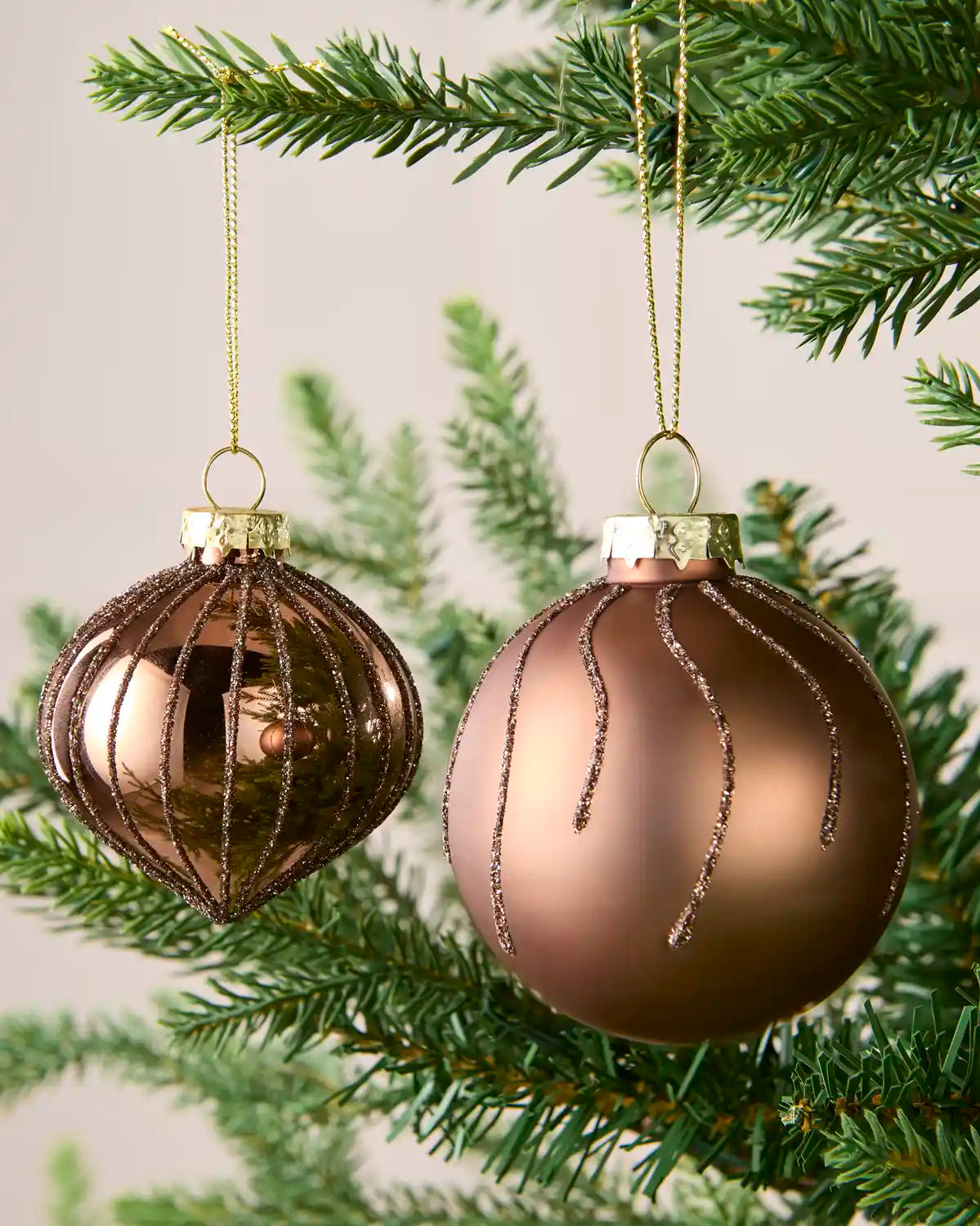 Two metallic Christmas tree ornaments hanging from a green pine branch: one ribbed Mocha ball with gold wire detailing and one smooth Mocha orb with dangling gold glitter lines, against a soft light background.
