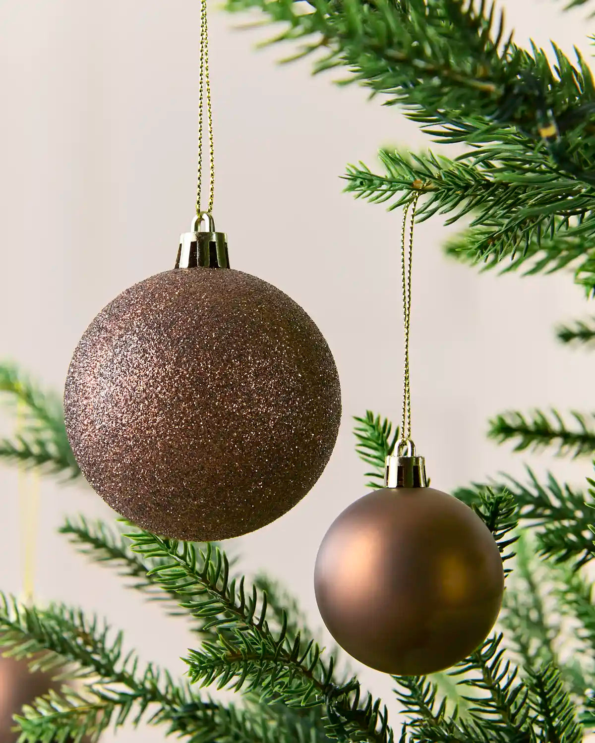 Two metallic Christmas tree ornaments hanging from a green pine branch against a soft light background. One ornament is large, glittery Mocha, and the other is smaller, smooth, and metallic Mocha.