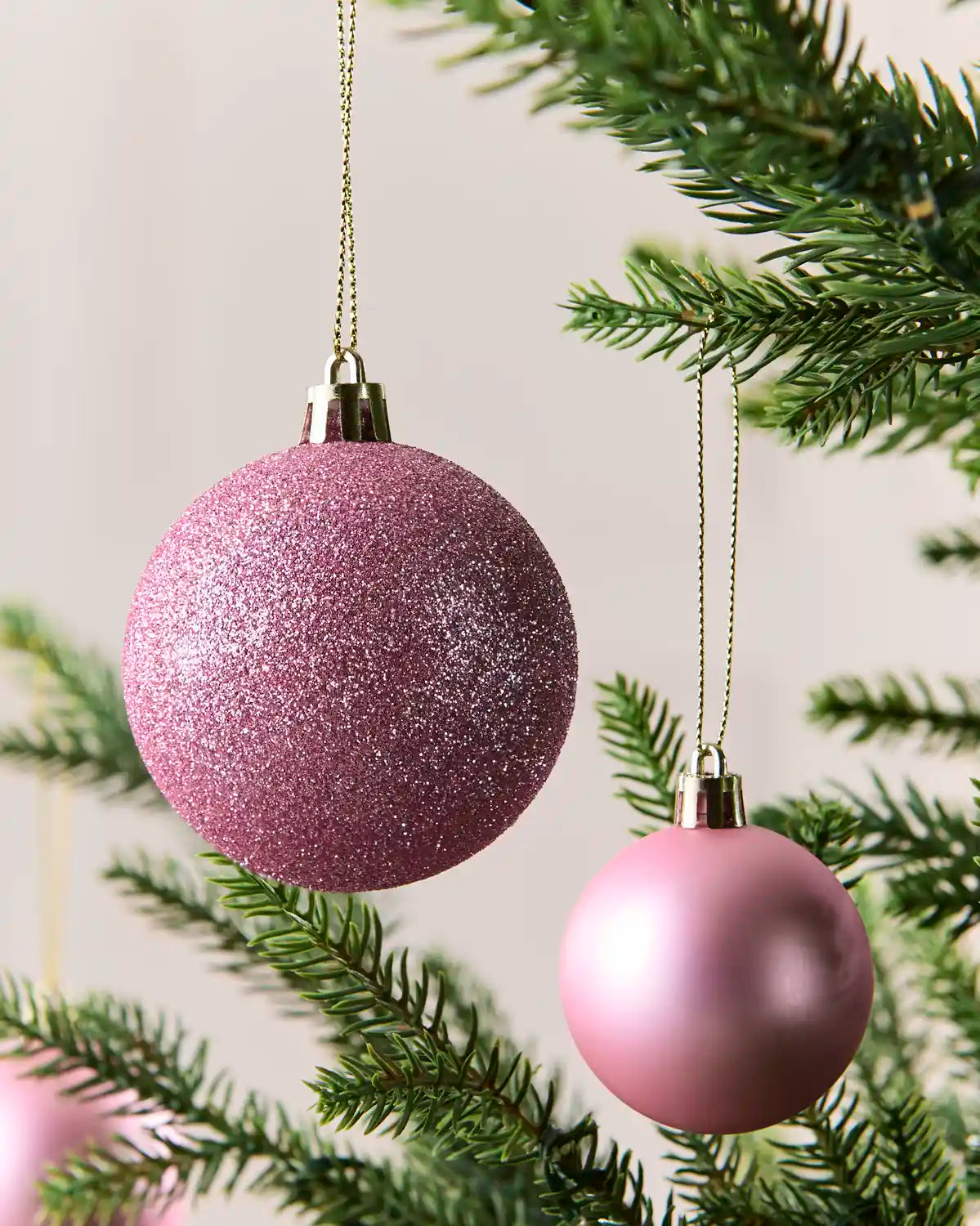Two pink Christmas tree ornaments hanging from a green pine tree branch against a soft light background.