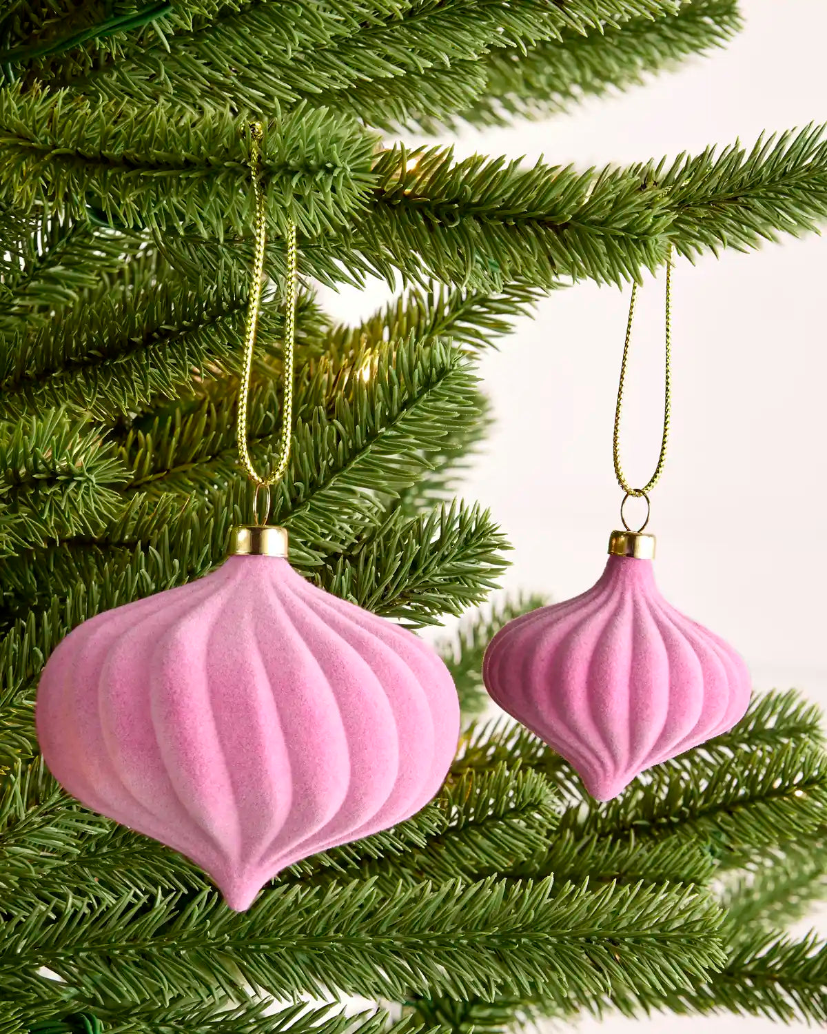 Two pink ribbed Christmas tree ornaments hanging from green pine branches with gold hanging cords.