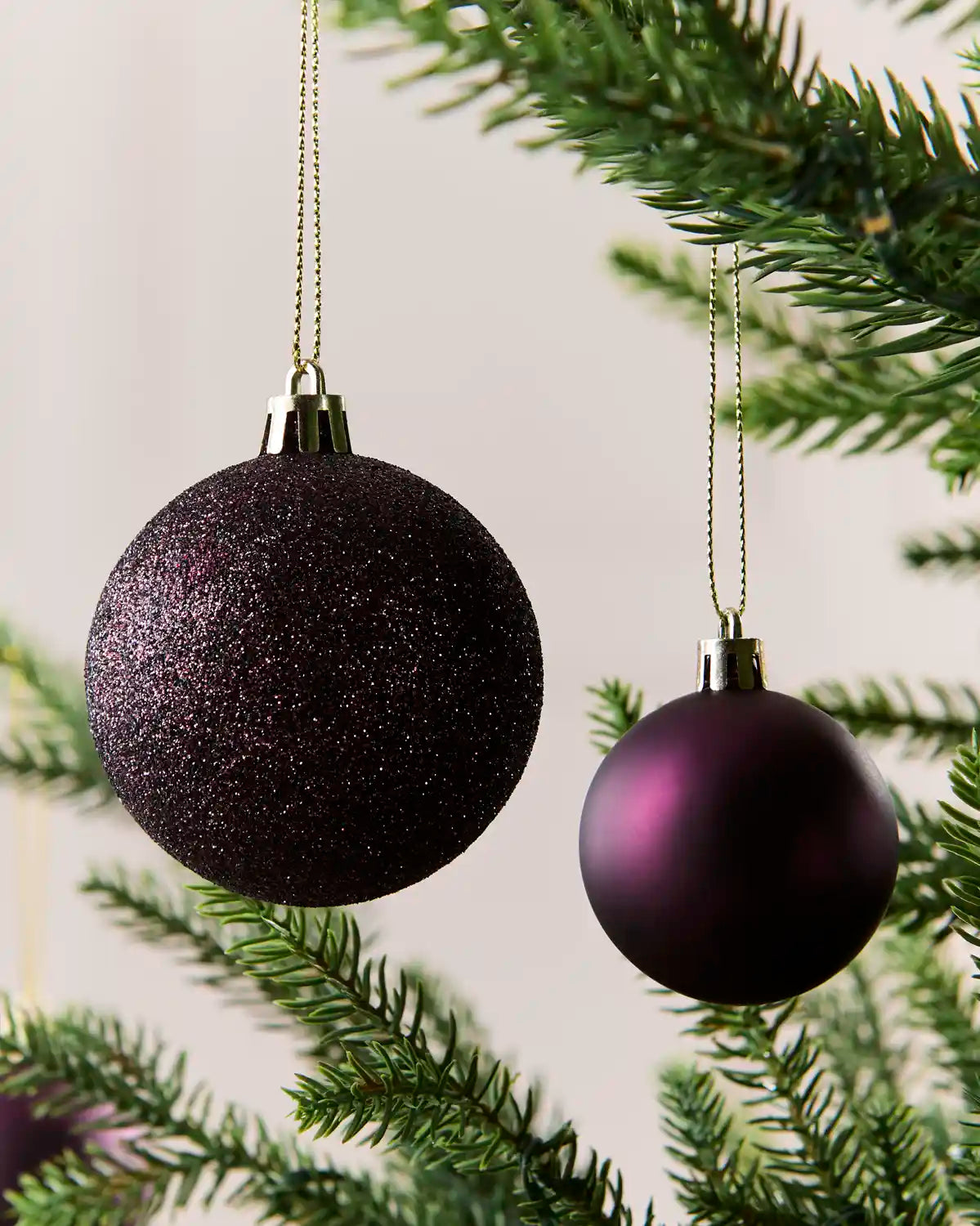 Two purple Christmas tree baubles hanging from green pine branches against a soft off-white background.