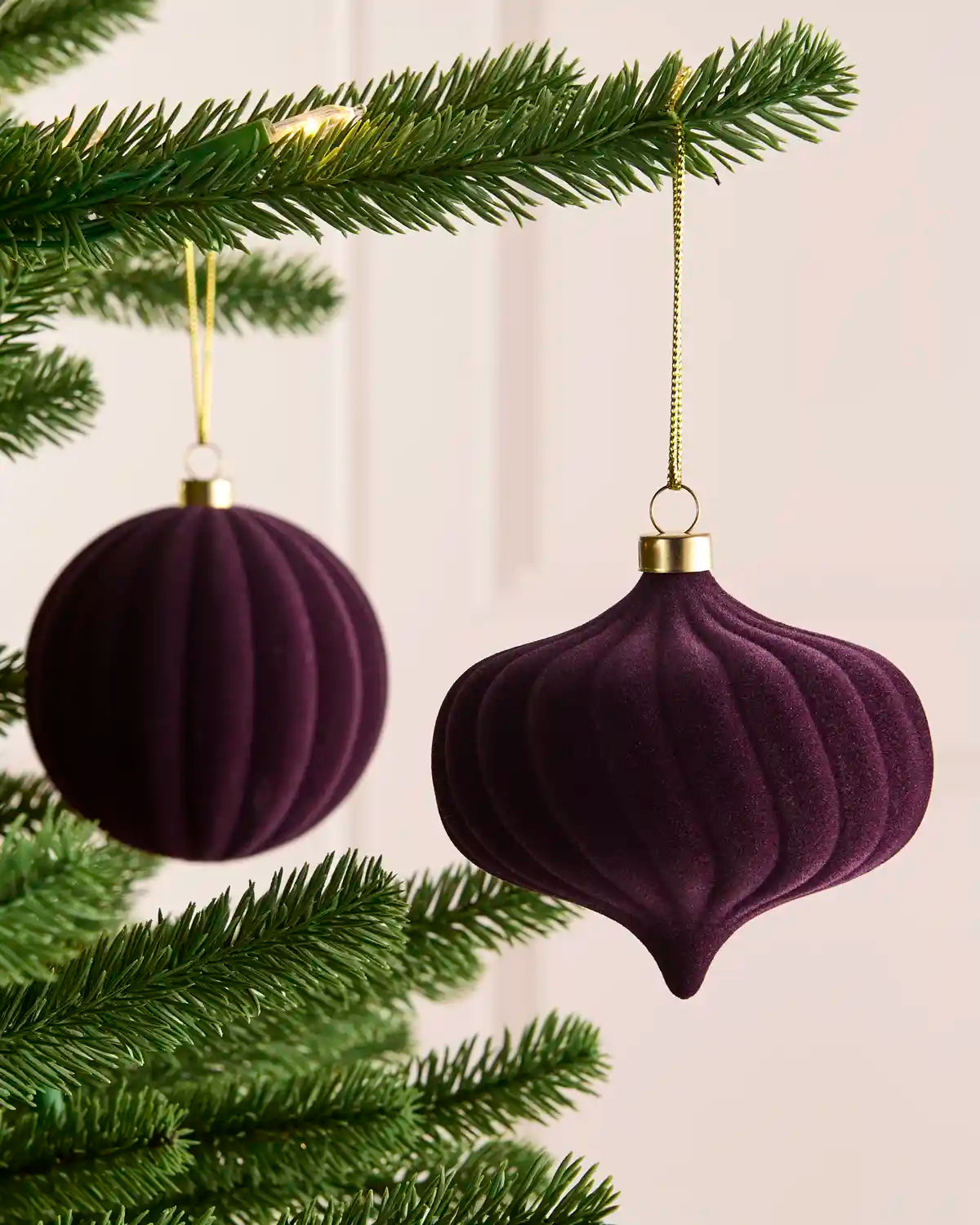 Two purple velvet Christmas tree ornaments hanging from a green pine tree branch against a soft cream background.