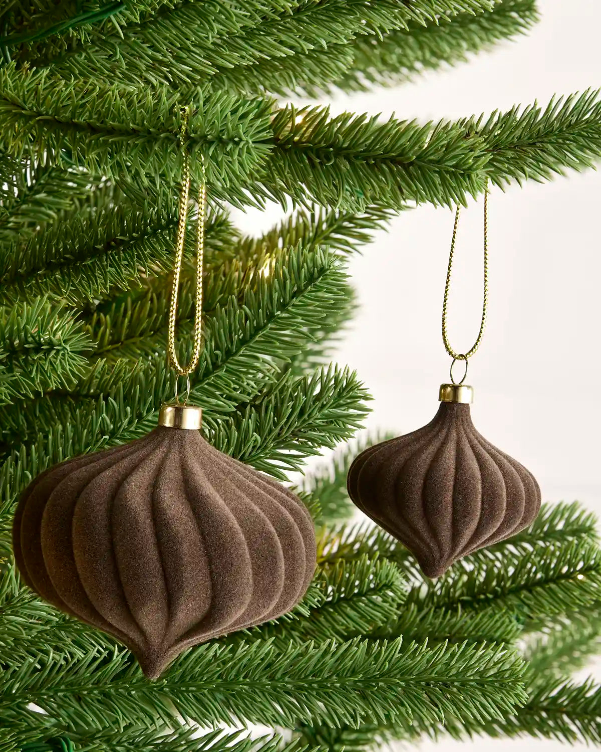 Two ribbed brown Christmas tree ornaments with gold hanging loops and caps, hanging from green pine tree branches against a light background.