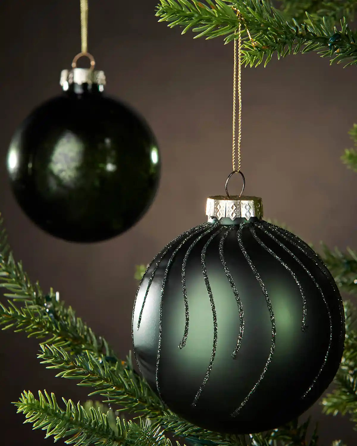 Two sage green Christmas ornaments hanging on a tree branch with a dark background