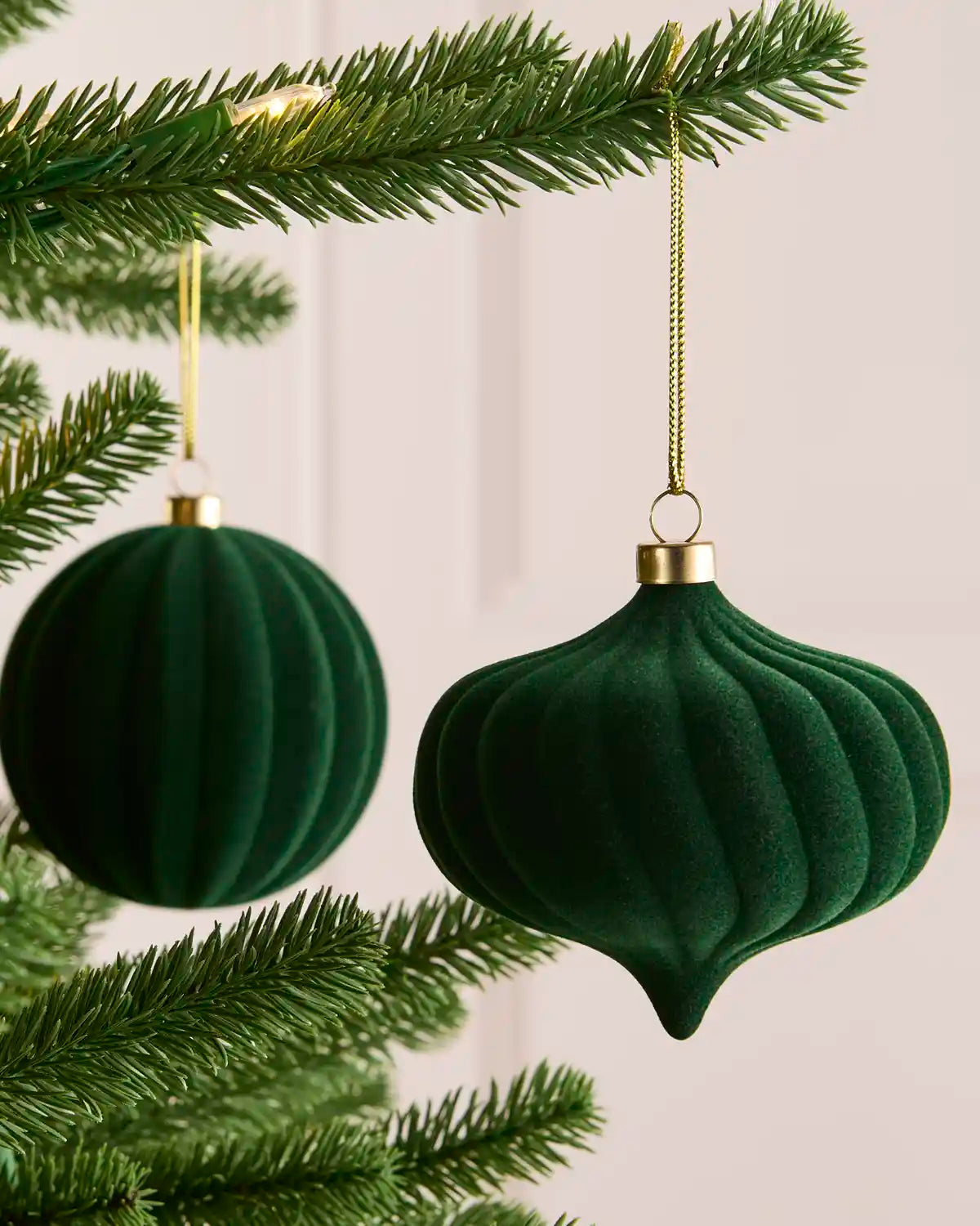 Two sage green ribbed velvet Christmas tree ornaments hanging from a pine tree branch against a soft light background.