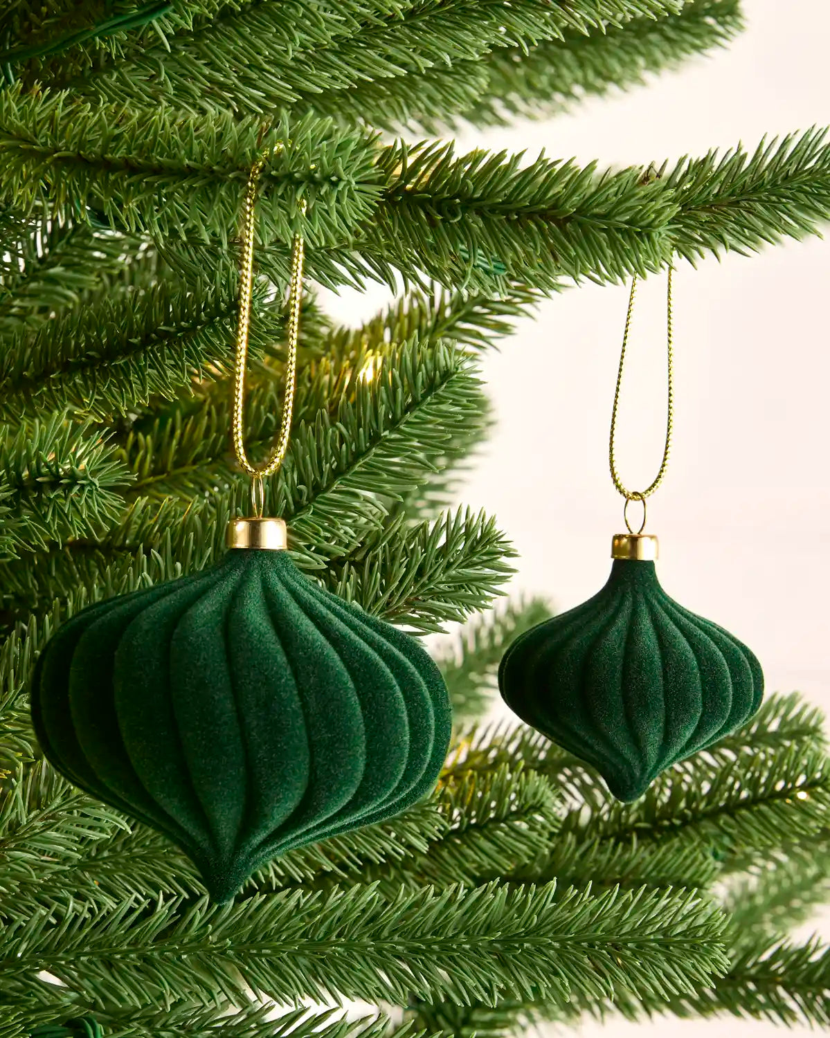 Two sage green ribbed velvet Christmas tree ornaments hanging from gold chains on green pine tree branches.