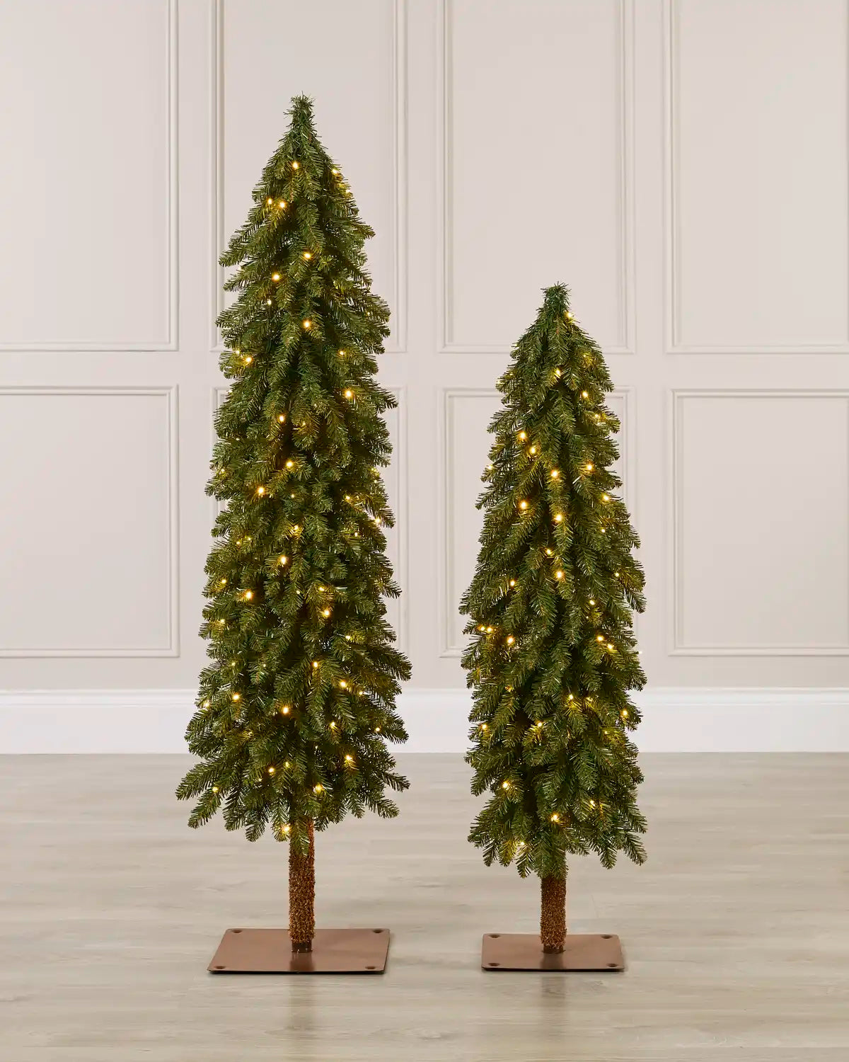 Two slim green Christmas trees with warm white lights, shown side by side on square brown bases against a white panelled wall