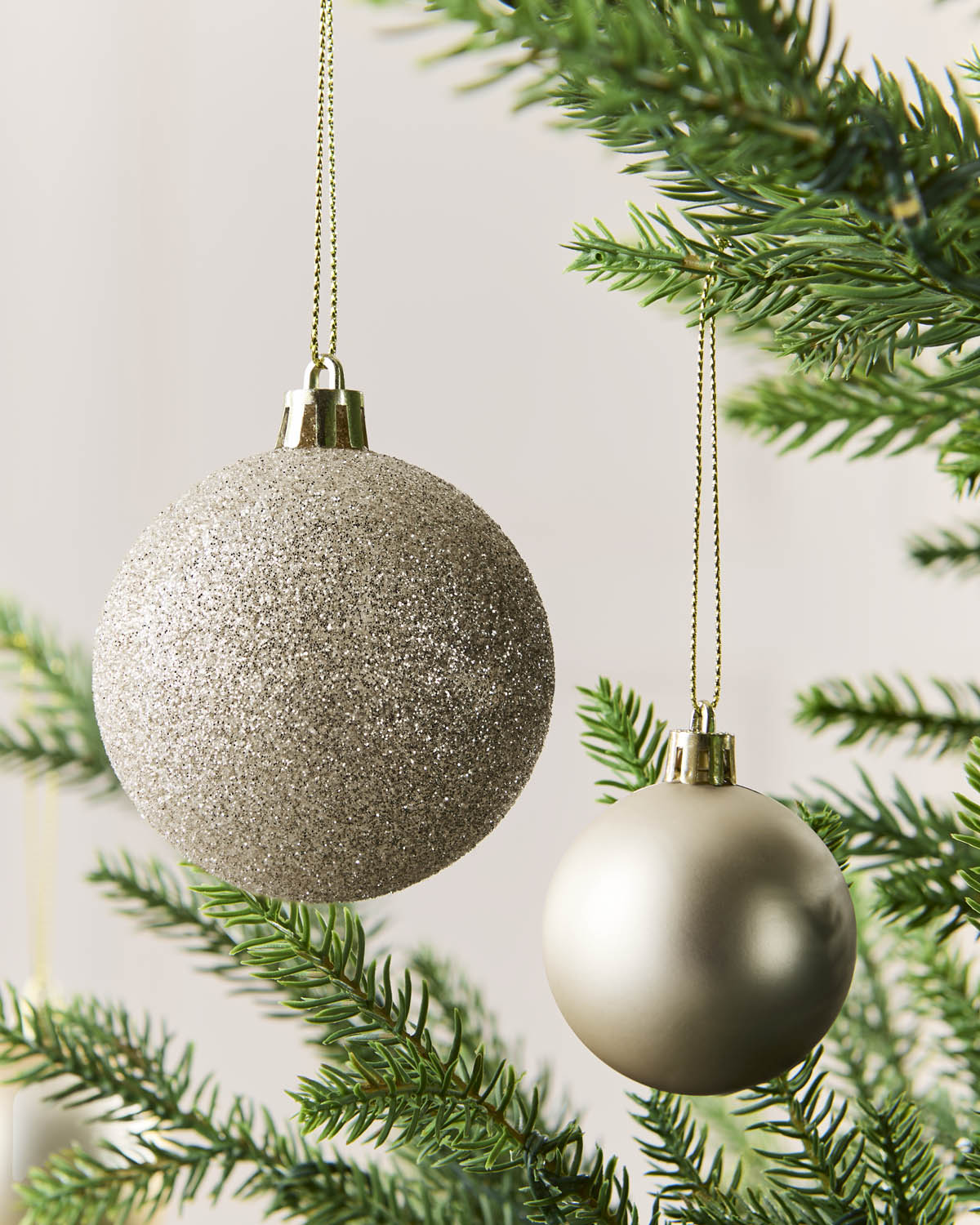 Two spherical Christmas tree ornaments hanging from a green pine branch. One ornament is large, textured, and glittery Latte, while the other is smaller, smooth, and metallic champagne gold.