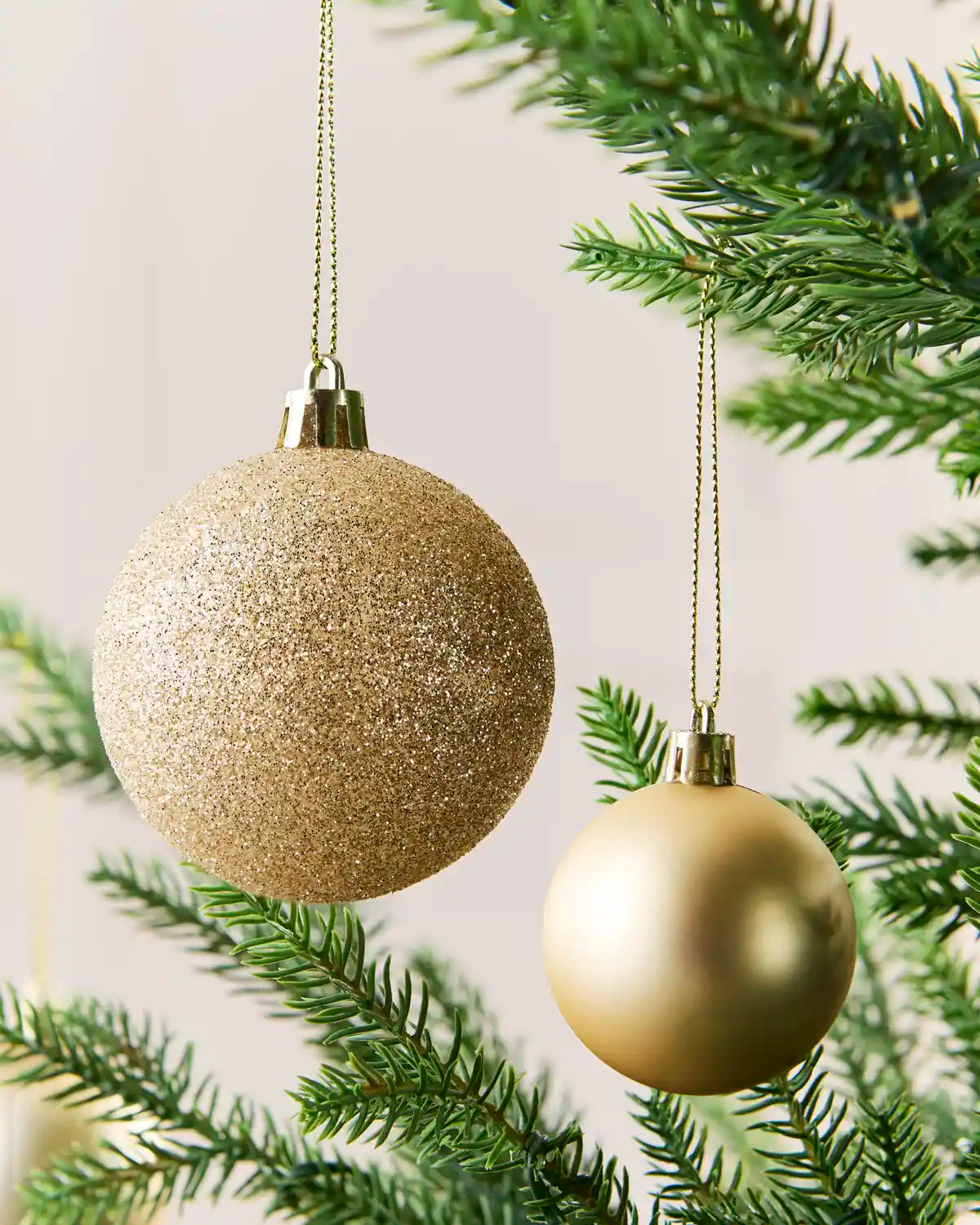 Two spherical Christmas tree ornaments hanging from a green pine branch. One ornament is large, textured, and glittery Latte, while the other is smaller, smooth, and metallic champagne gold.
