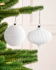 Two white Christmas tree ornaments hanging from a green pine tree branch against a soft cream wall background.