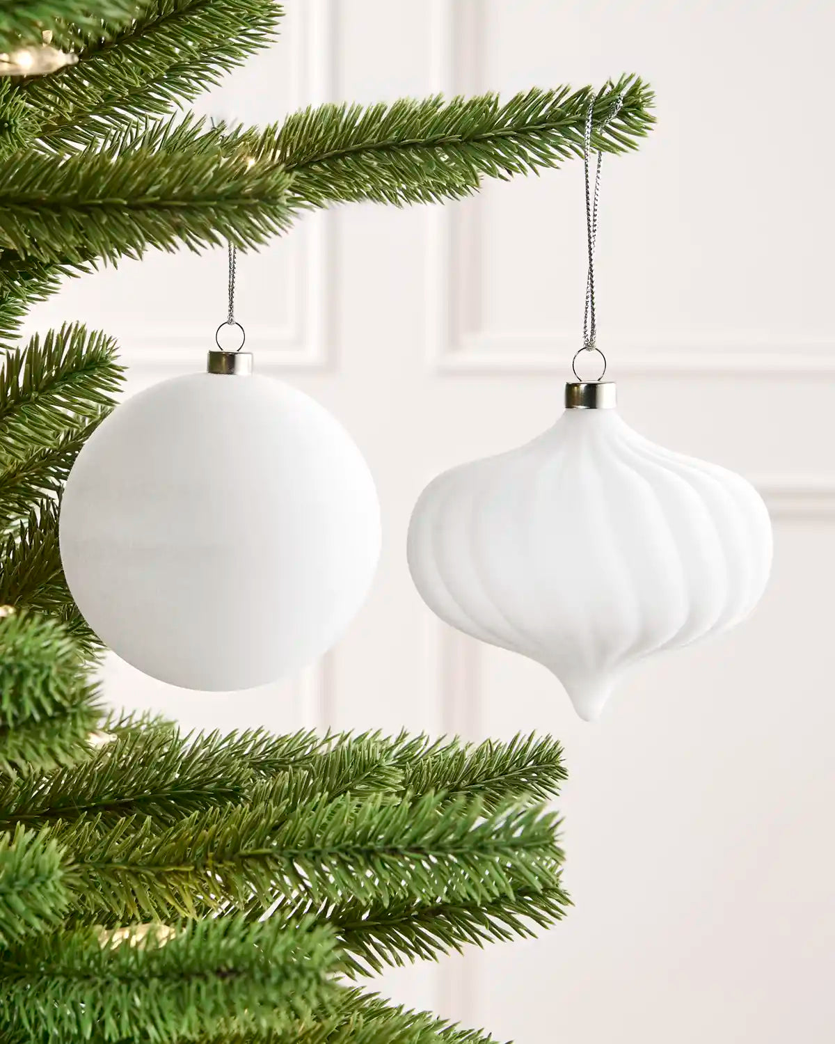 Two white Christmas tree ornaments hanging from green pine branches against a soft light pink wall.
