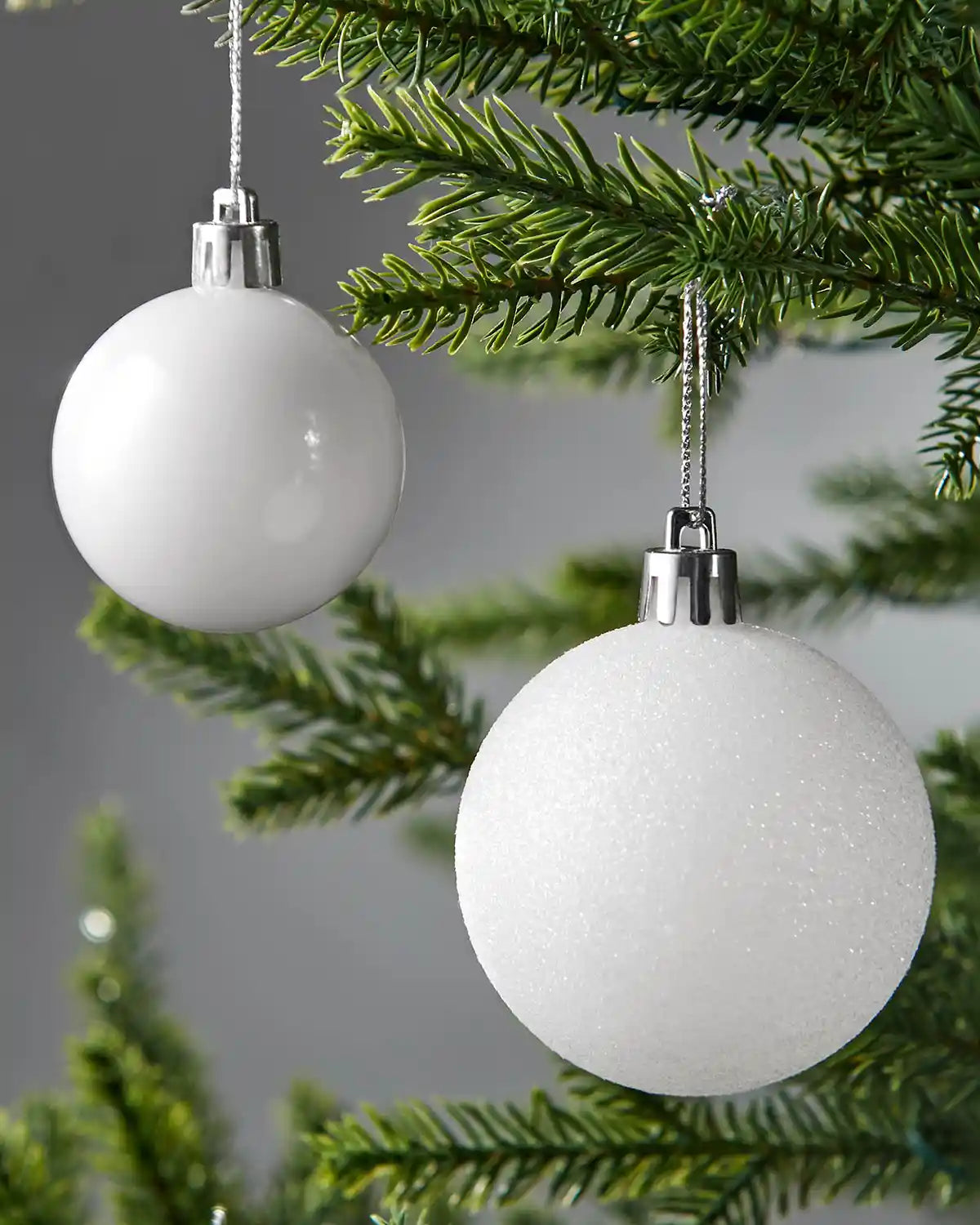 Two white baubles with silver tops hanging on a Christmas tree.