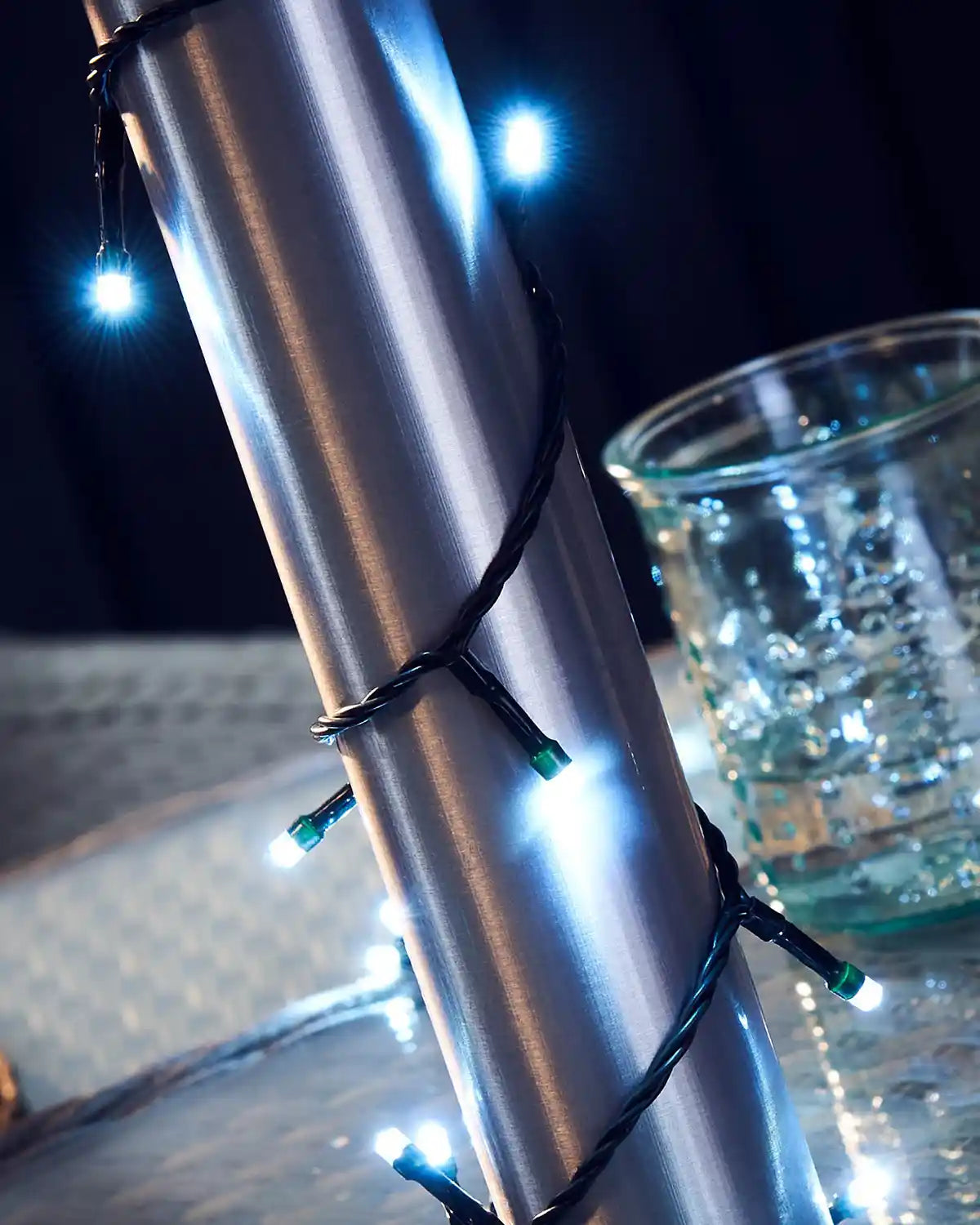 Umbrella post wrapped in ice white fairy lights on a grey wicker table and glass in background 