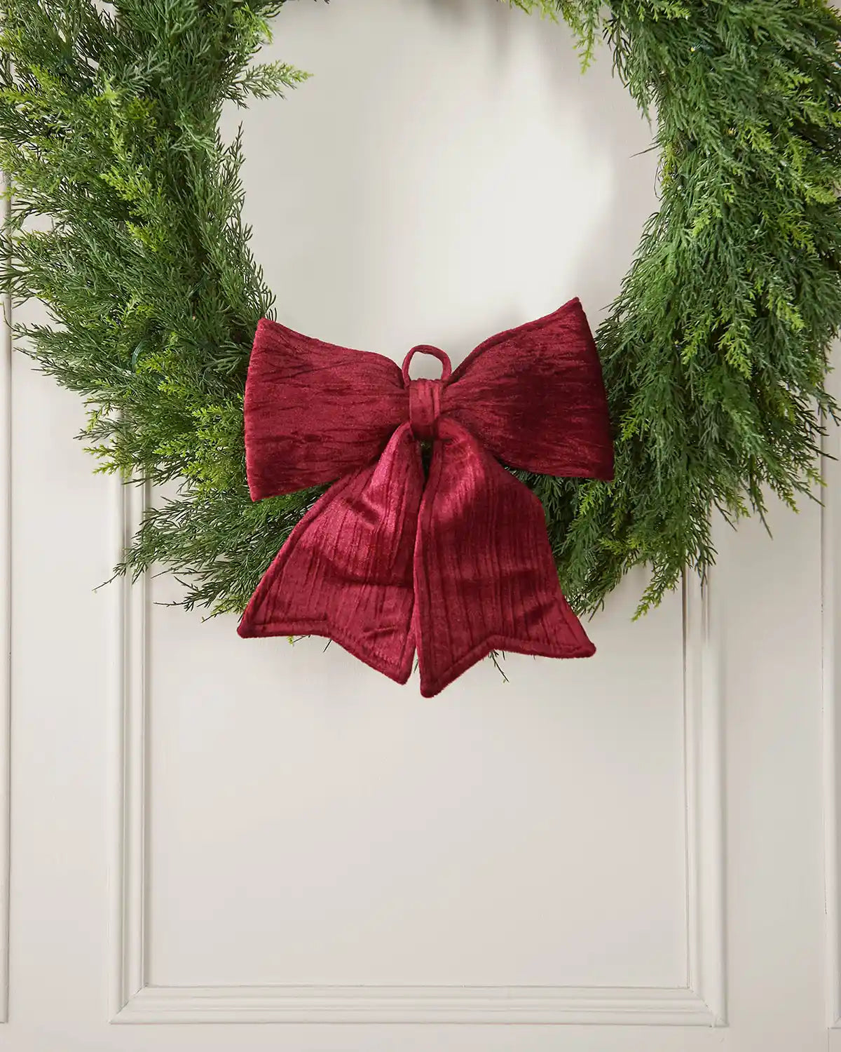 Vintage-style burgundy velvet bow placed on the bottom of a green wreath hanging on a white door
