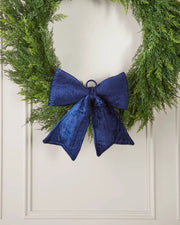 Vintage-style navy blue velvet bow attached to bottom of green wreath in front of a white background