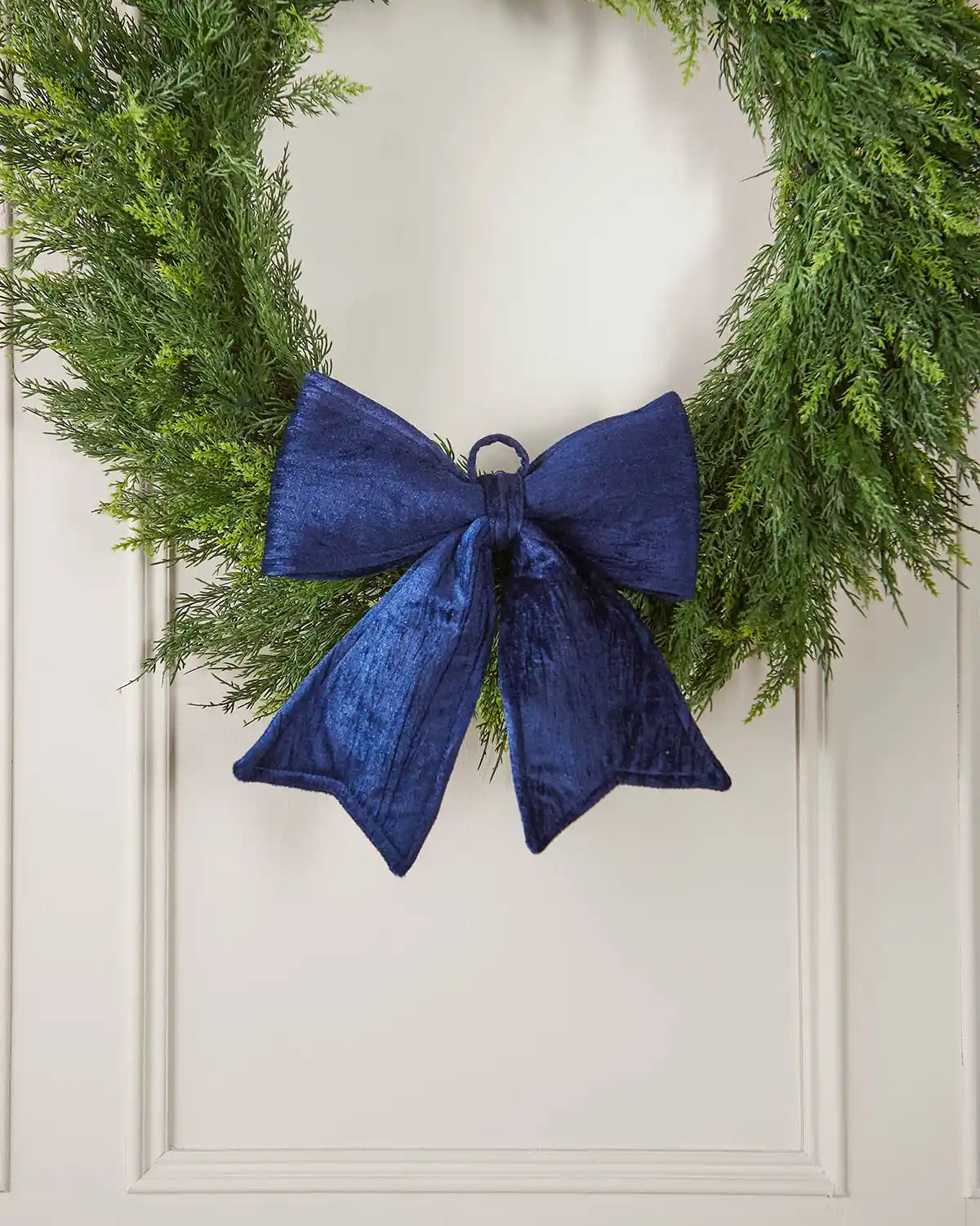 Vintage-style navy blue velvet bow attached to bottom of green wreath in front of a white background