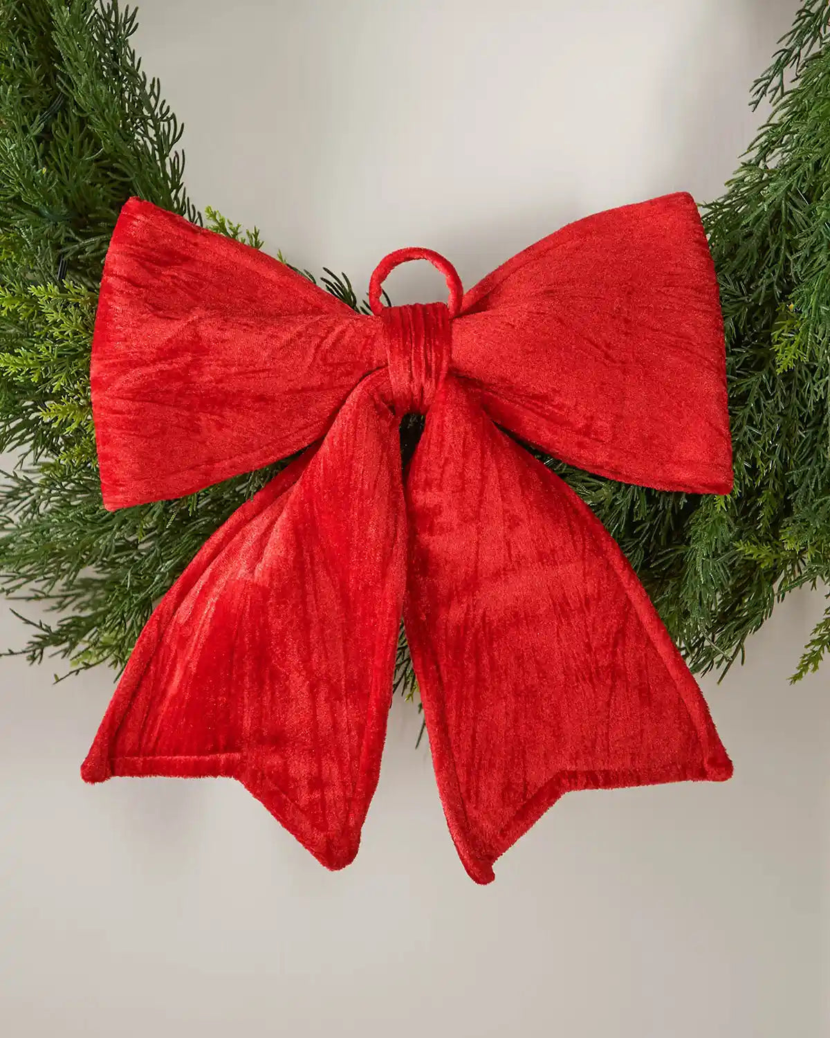 Vintage-style red velvet bow pinned on the bottom of a green wreath, in front of a background of a white door