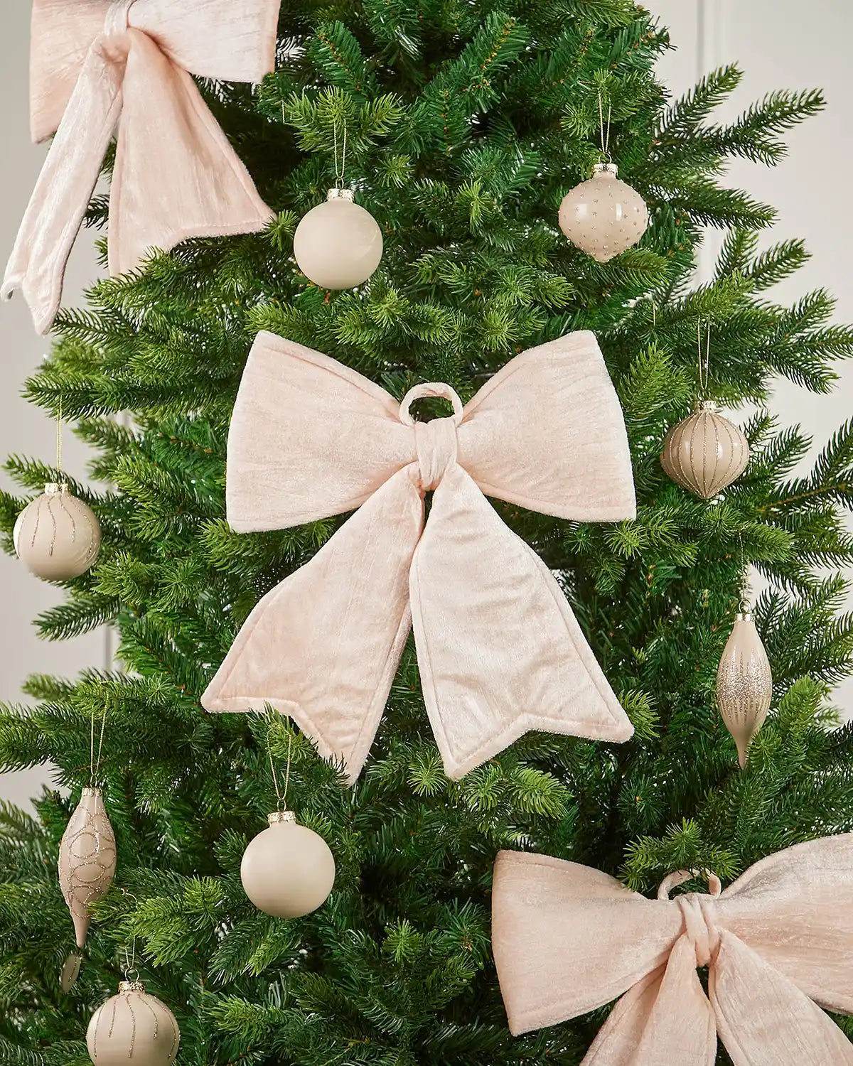 Vintage-style cream velvet Christmas bow attached to the centre of a tree surrounded by identical bows and cream baubles in front of white panelling