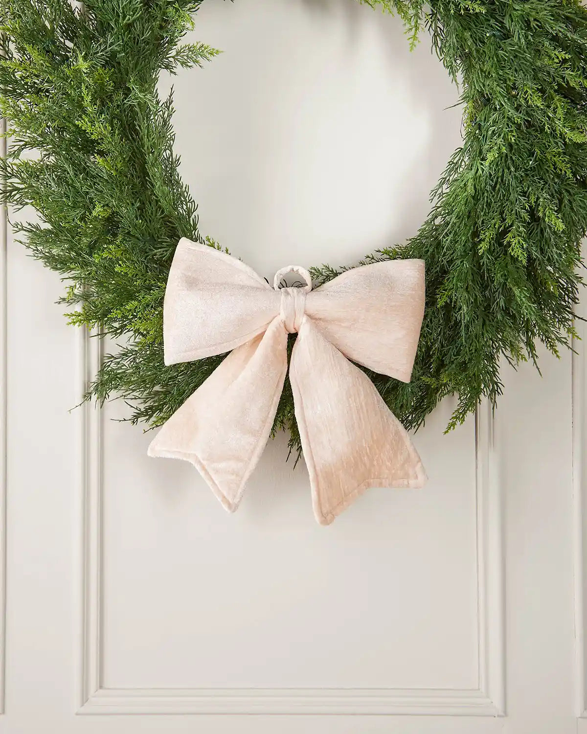 Vintage-style cream velvet Christmas bow attached to the bottom of a green wreath in front of white panelling