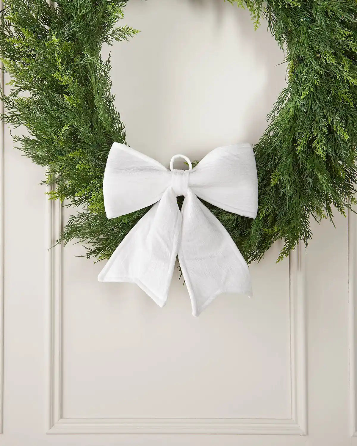 Vintage-style white velvet bow attached to bottom of wreath in front of a white background