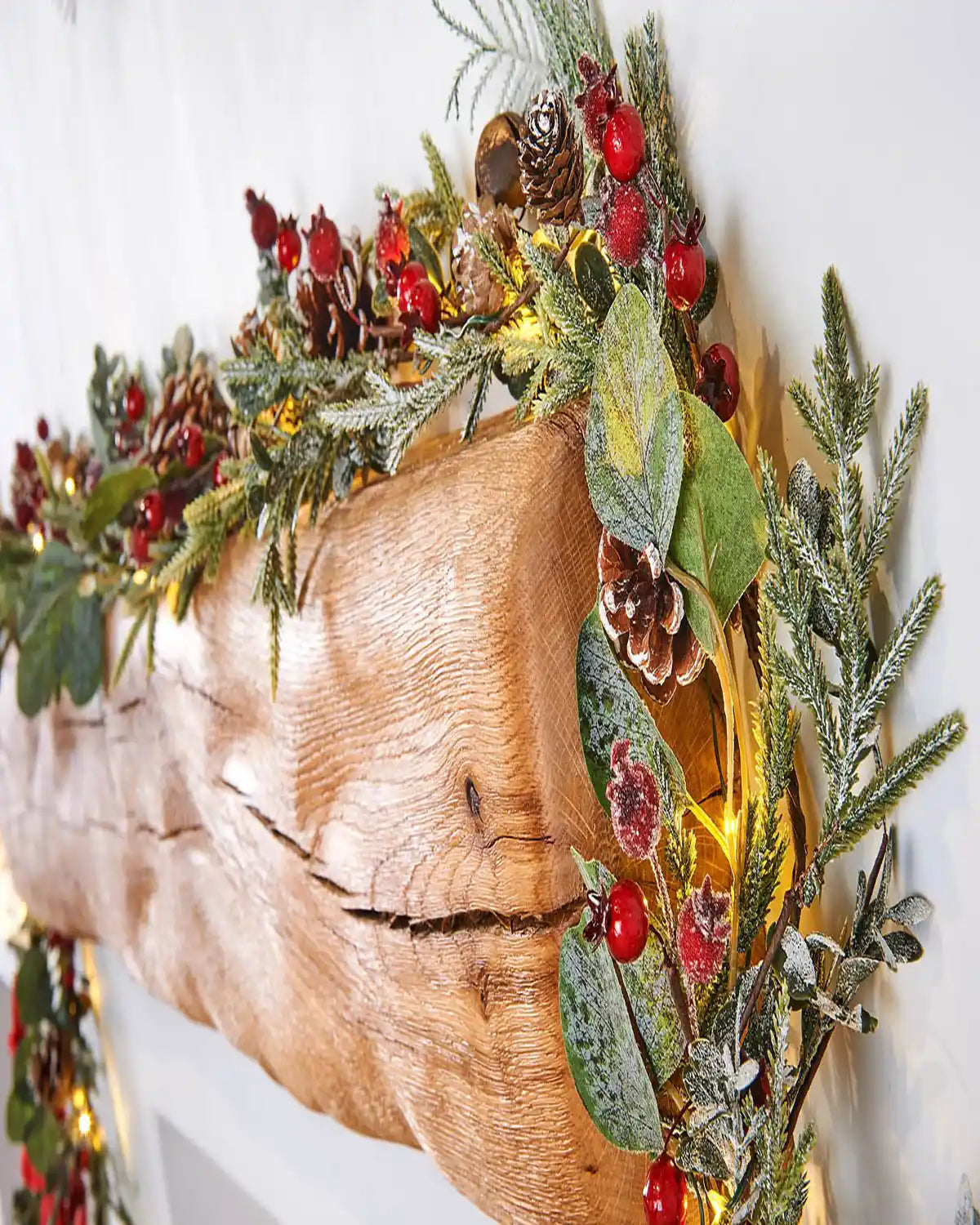 Close up decorative Christmas garland with Red berried, pinecones with lights on a wooden mantel against a white wall.