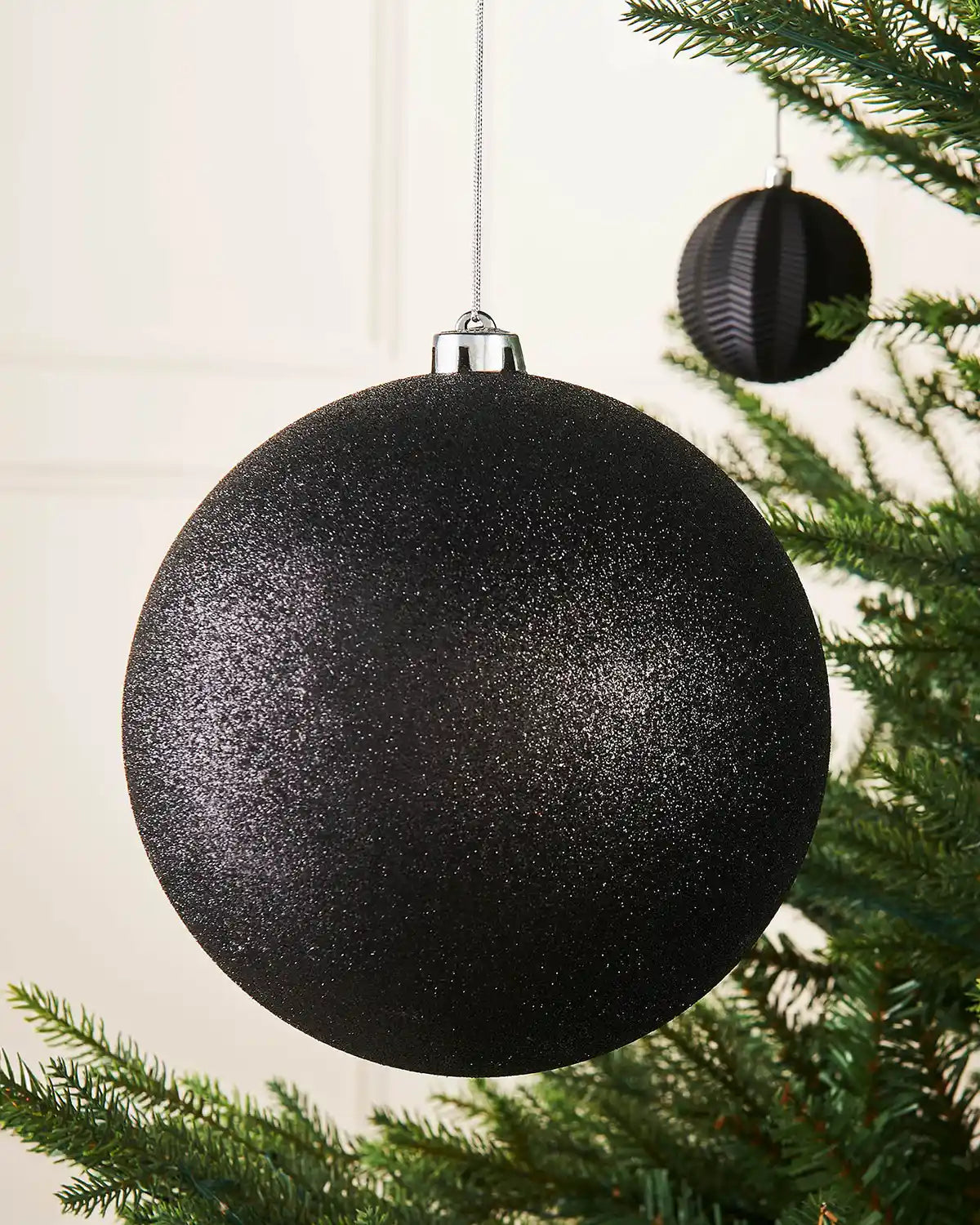 Black glittery oversized Christmas bauble on a Christmas tree branch with a smaller black bauble in background