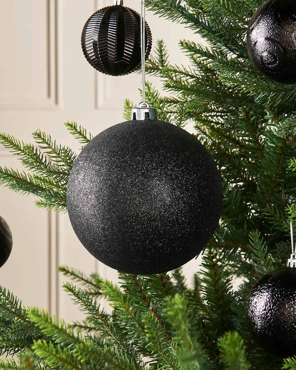 Black glitter oversized 15cm bauble on a Christmas tree branch with 4 smaller black baubles in background