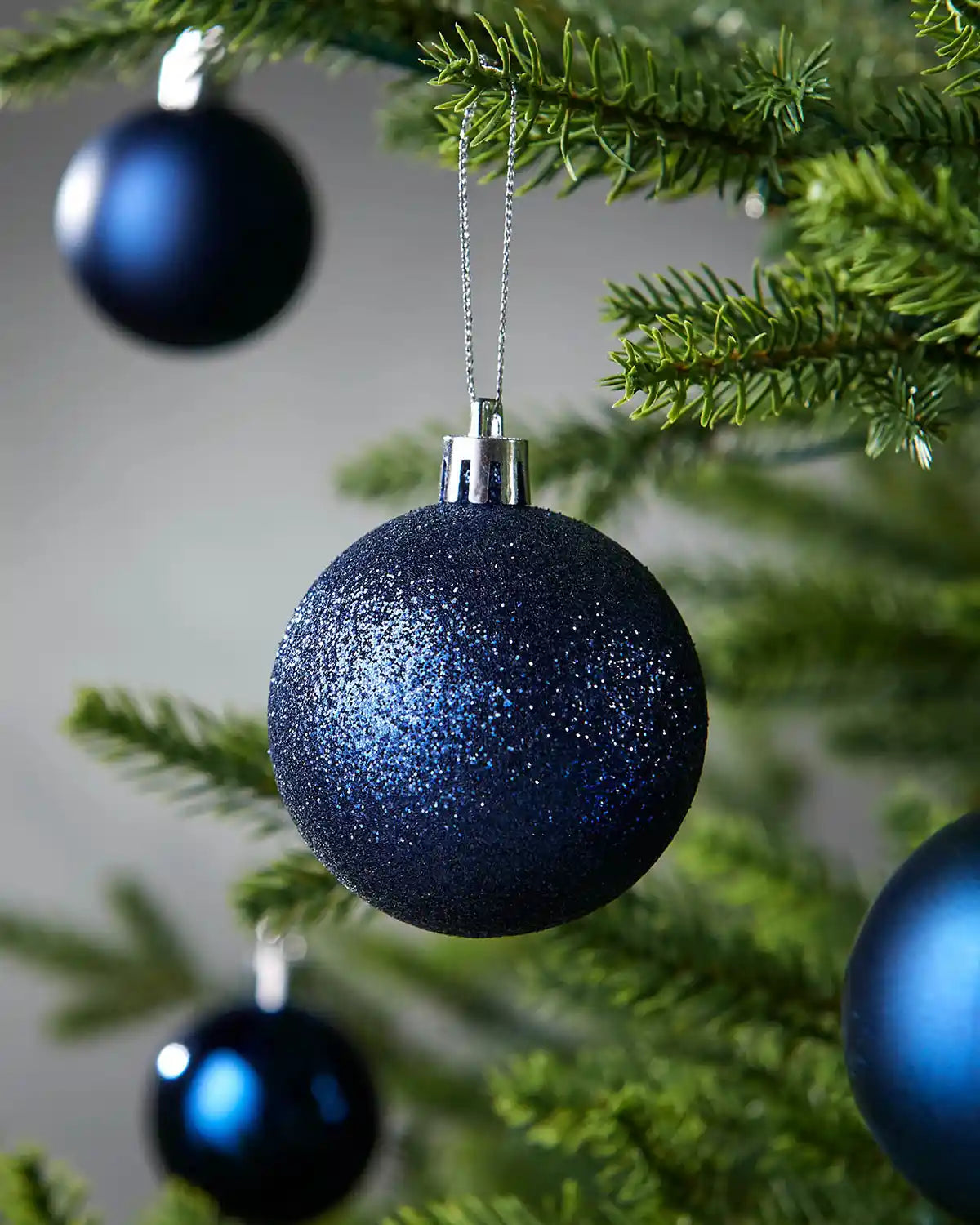 Blue glittery Christmas ball baubles hanging on a tree branch with a blurry grey background