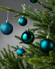 Teal Christmas tree ornaments hanging on green pine branches against a soft grey background.
