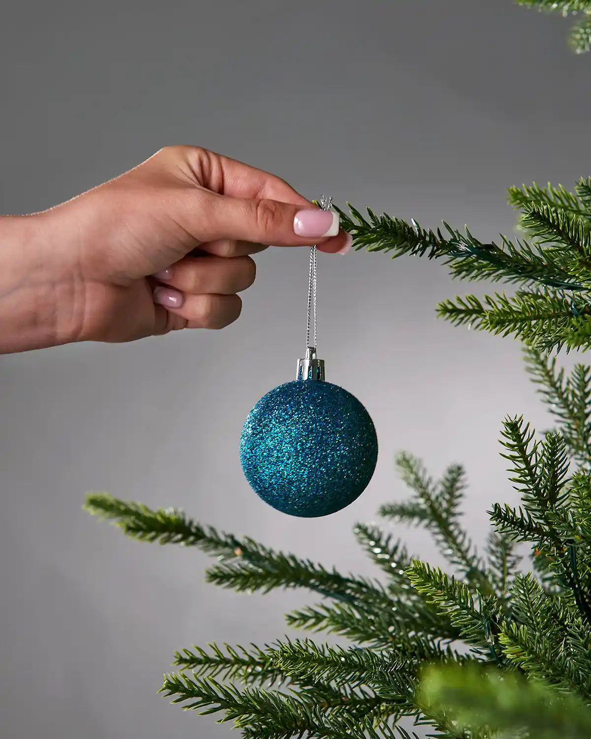 A hand with manicured nails hangs a glittery teal blue Christmas ornament from a green artificial pine tree branch against a neutral grey background.