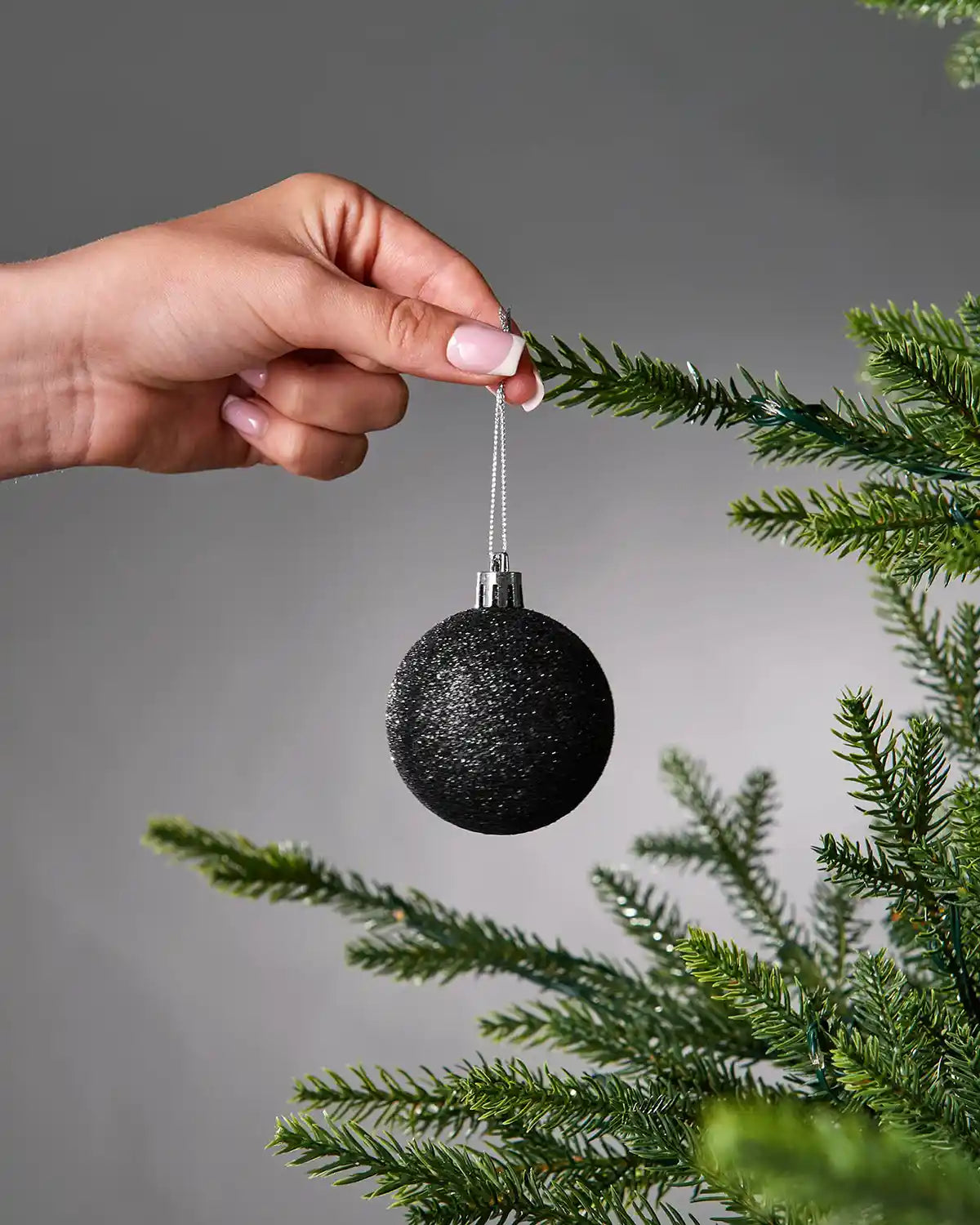 Hand hanging a black bauble on a Christmas tree branch against a grey background